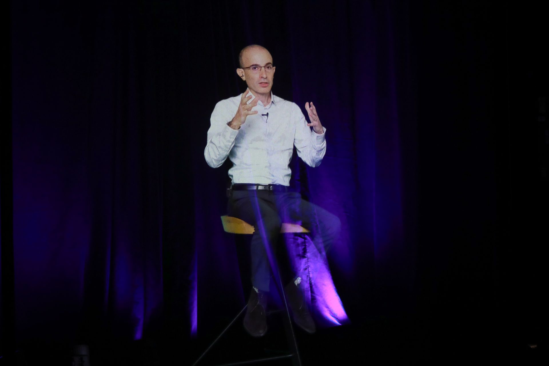 Man sits on a stool, gesturing with hands; stage with purple lighting in background.