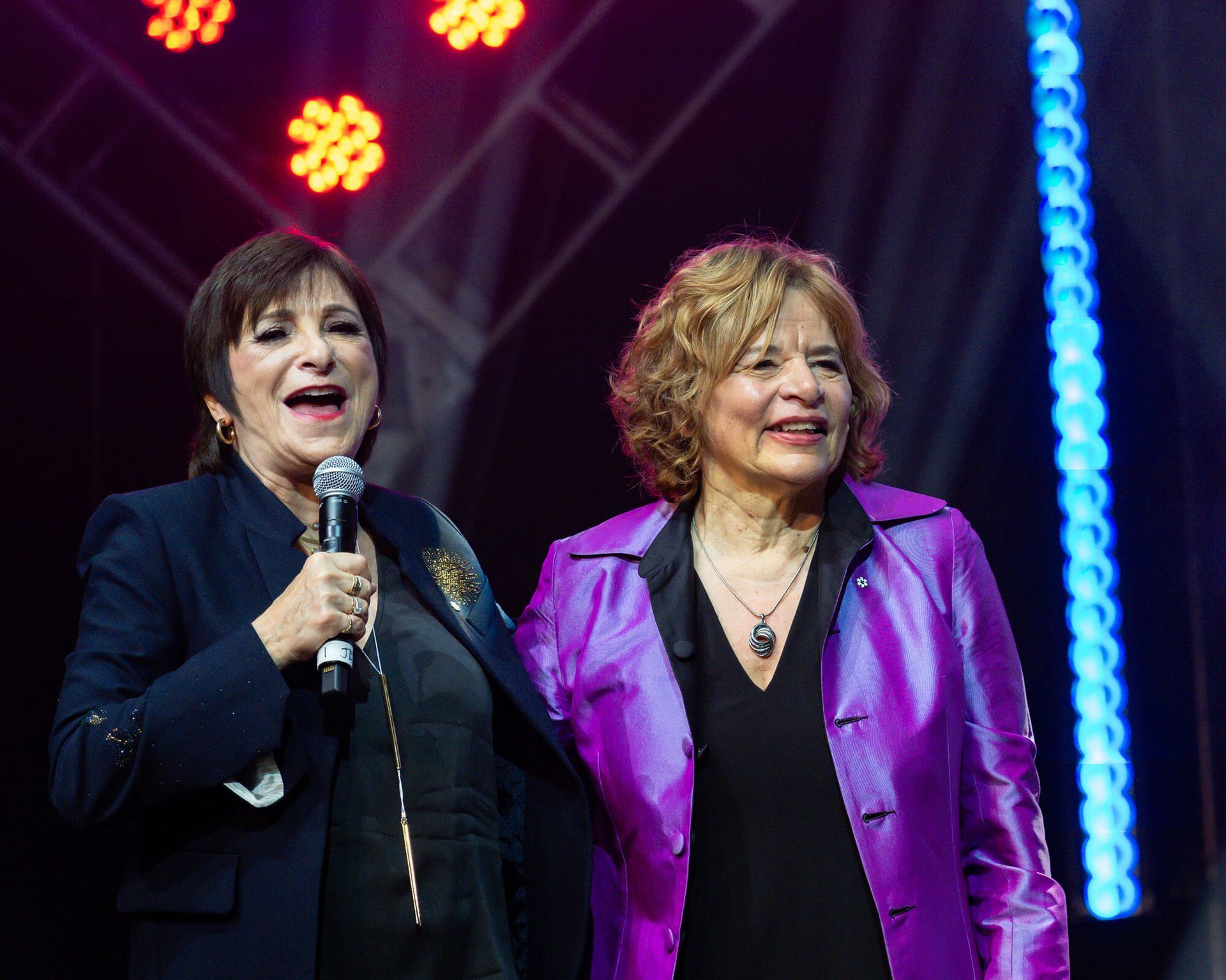 Two women are standing next to each other on a stage and one is holding a microphone.