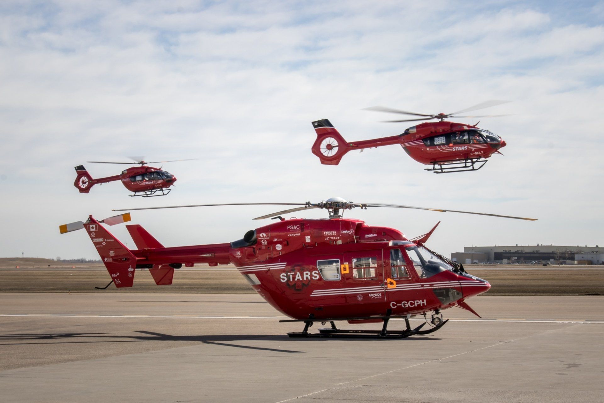 Three red helicopters are flying in formation on a runway.