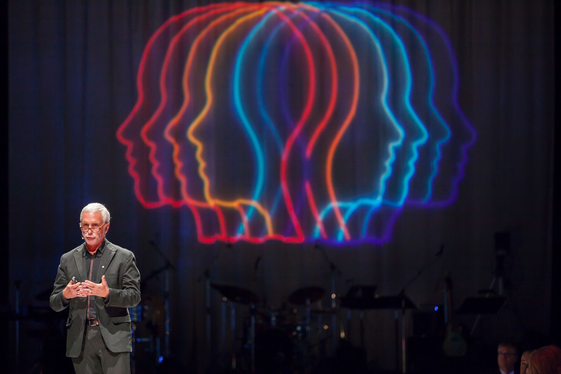 A man in a suit stands in front of a projection of faces
