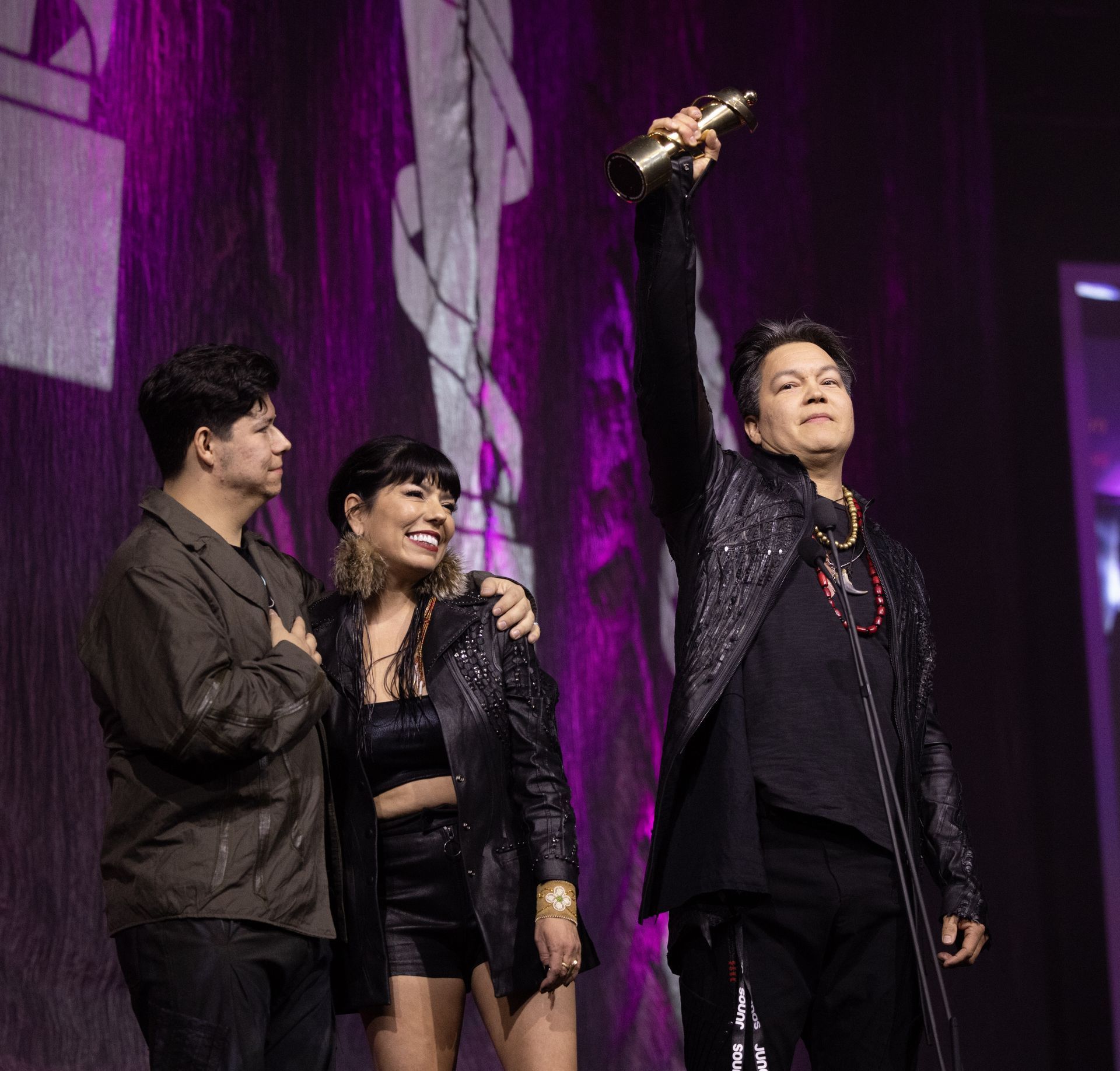 Man holding award on stage with two people; purple backdrop.