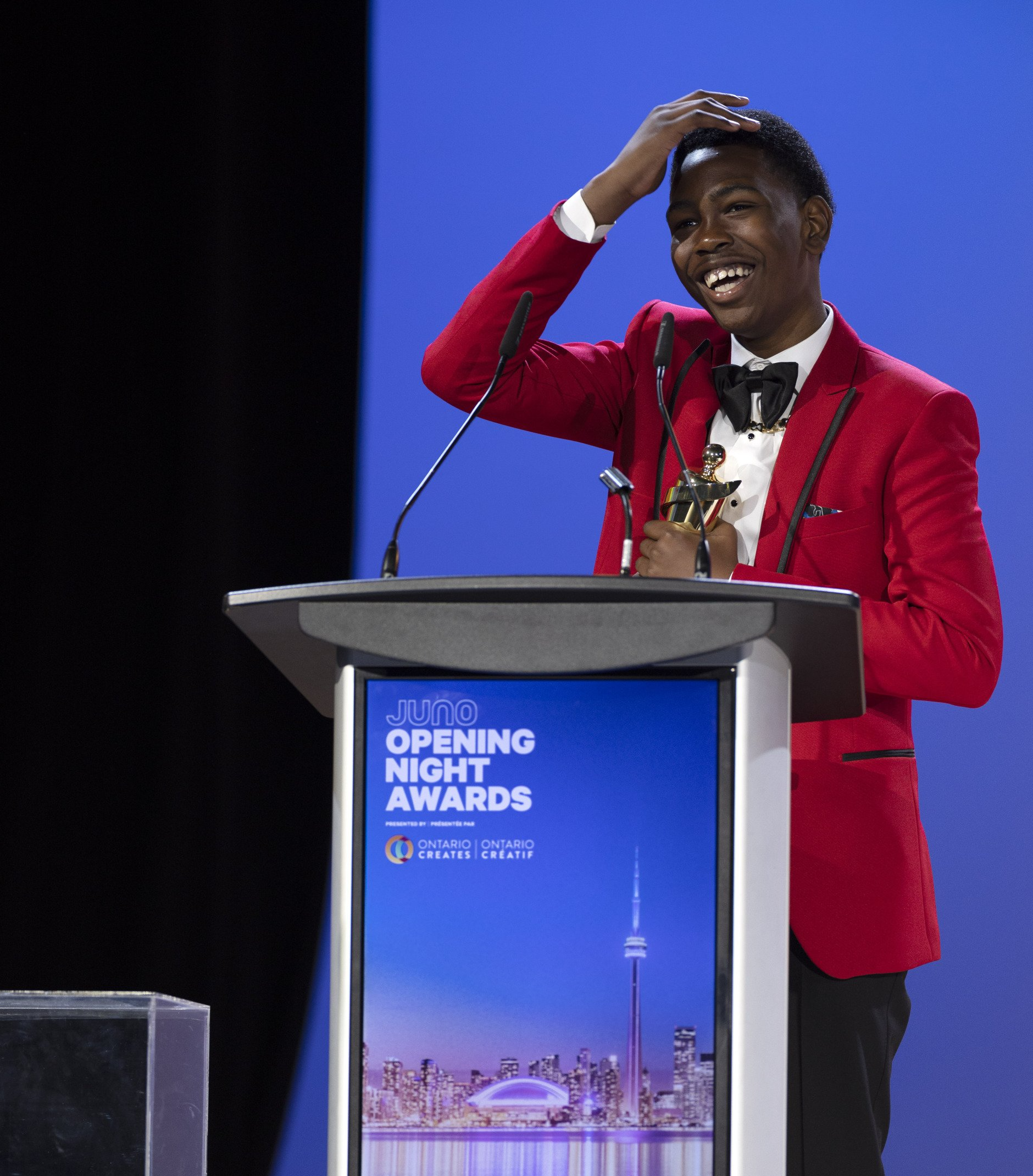 A man in a red jacket stands behind a podium that says the opening night awards