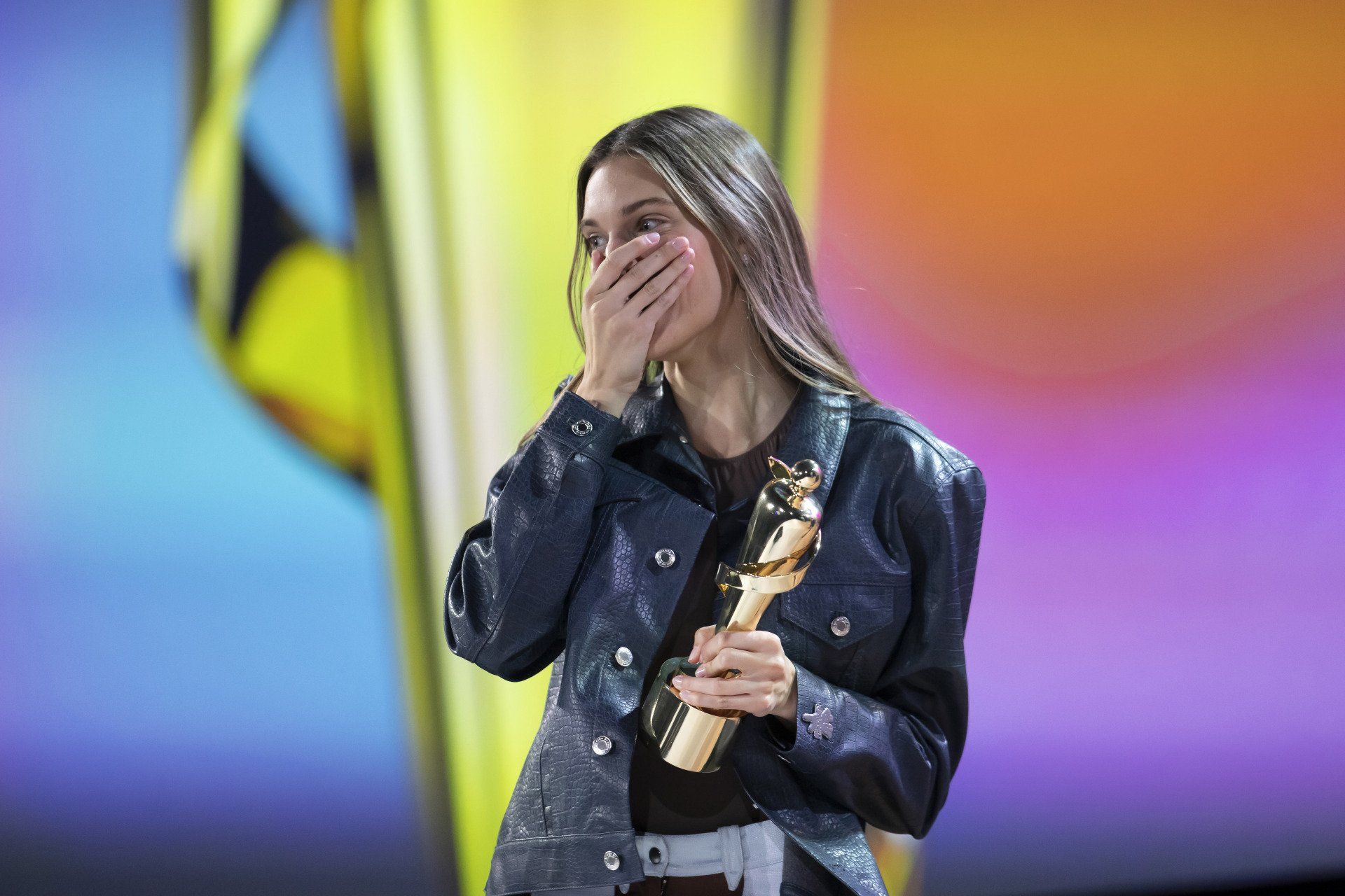 A woman is covering her mouth while holding a trophy on a stage.