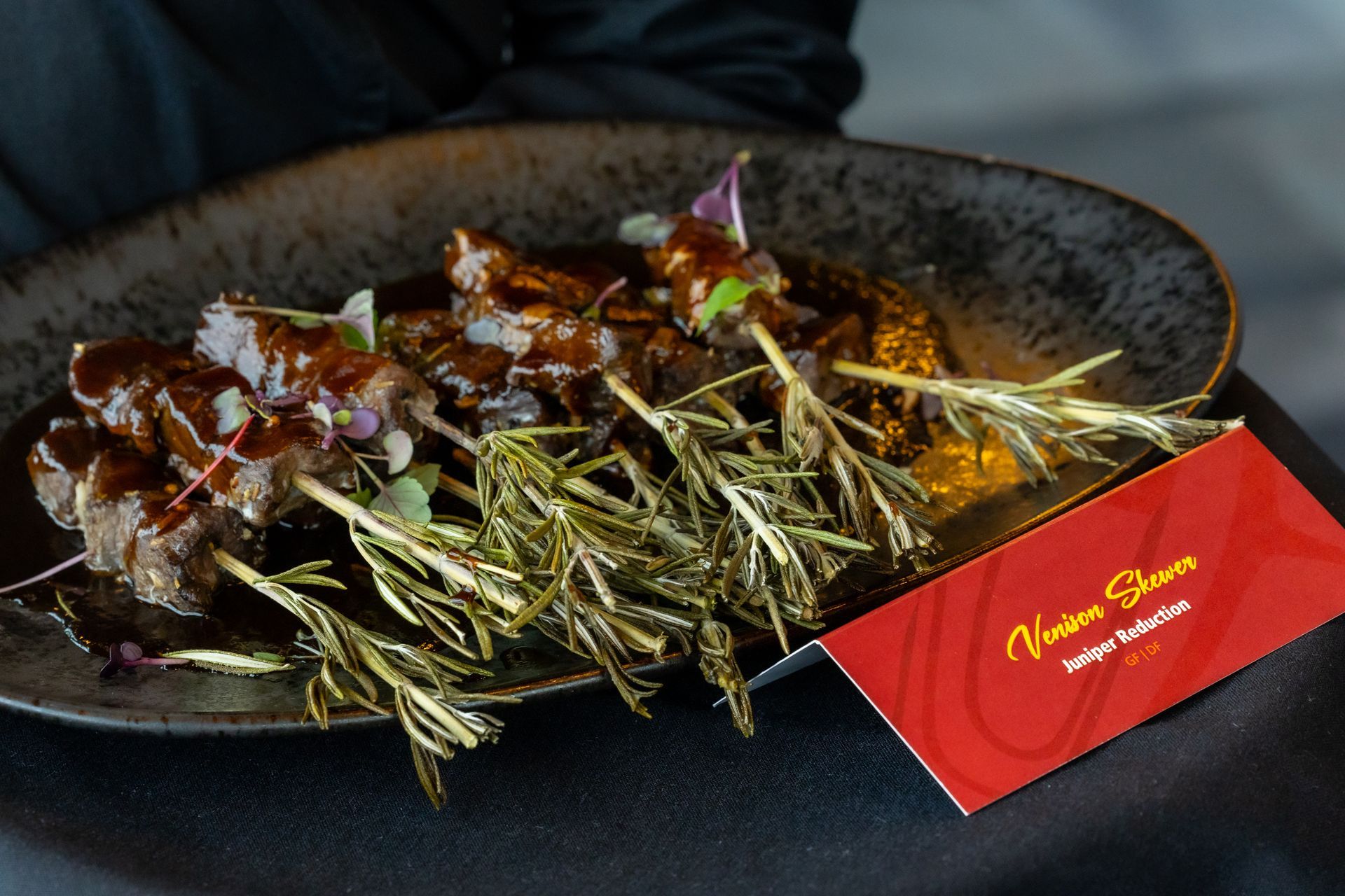 Skewered beef, rosemary garnish, drizzled sauce, on a dark plate, red card.