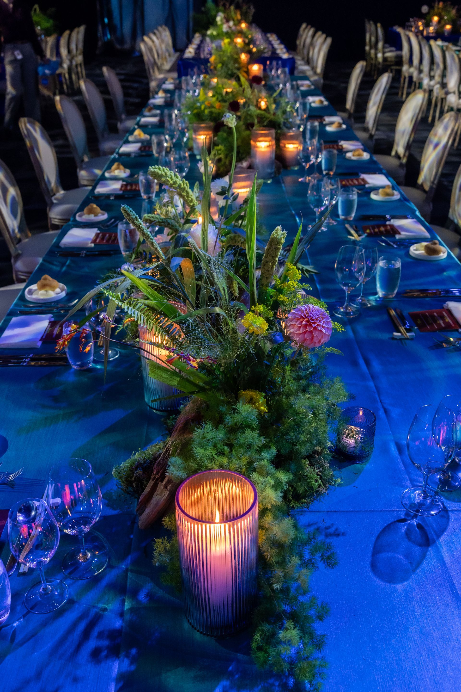 Long dining table set for an event with blue tablecloth, floral arrangements, and candles.
