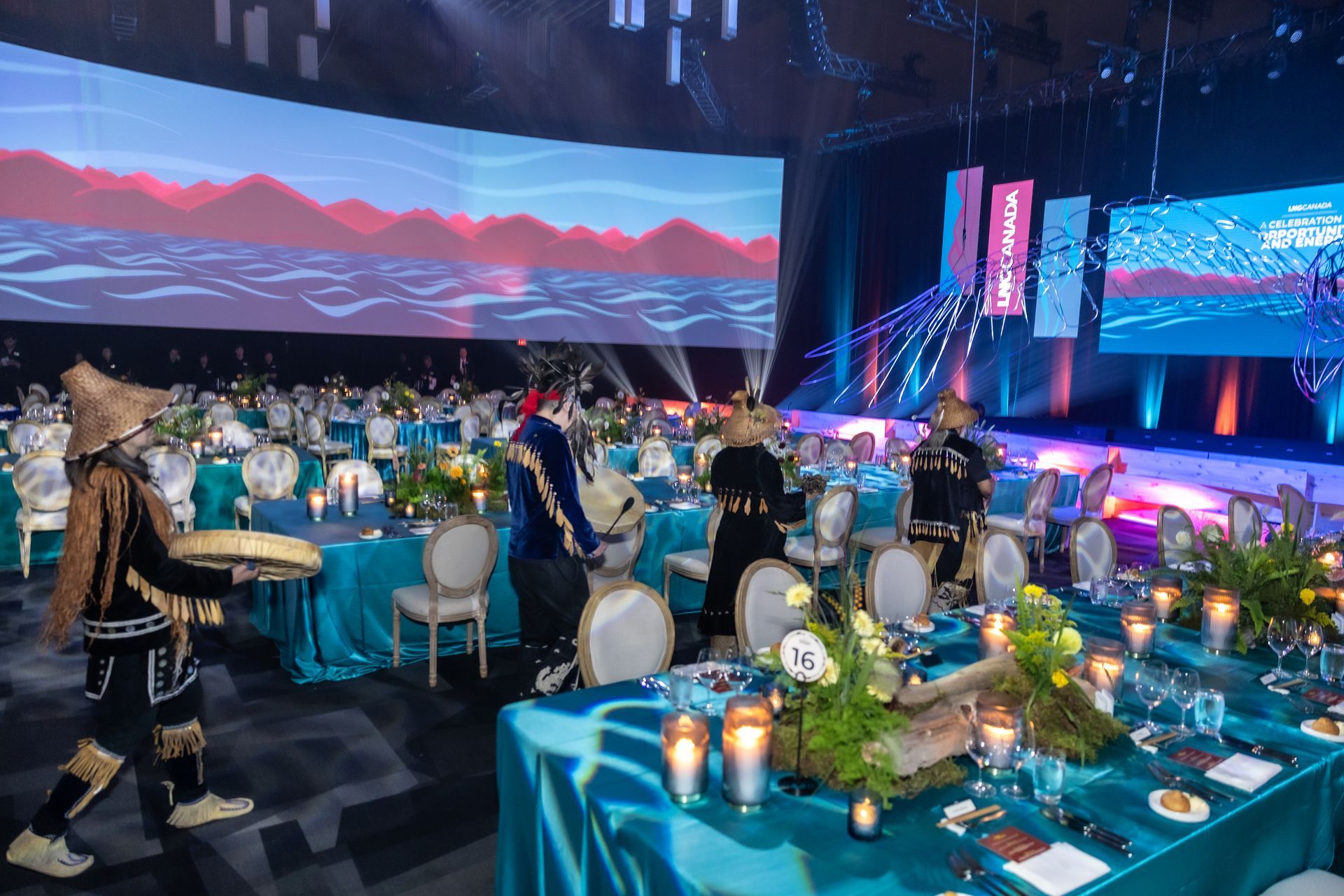 Indigenous dancers perform at a gala. Tables are set with teal linens, flowers, and candles.