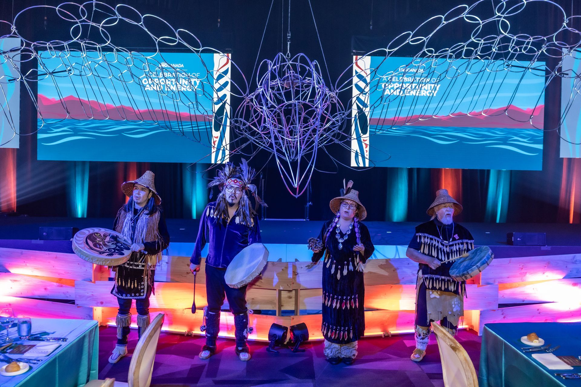 Group of four performers on stage, drumming and singing. Backdrop with screens and colorful lighting.