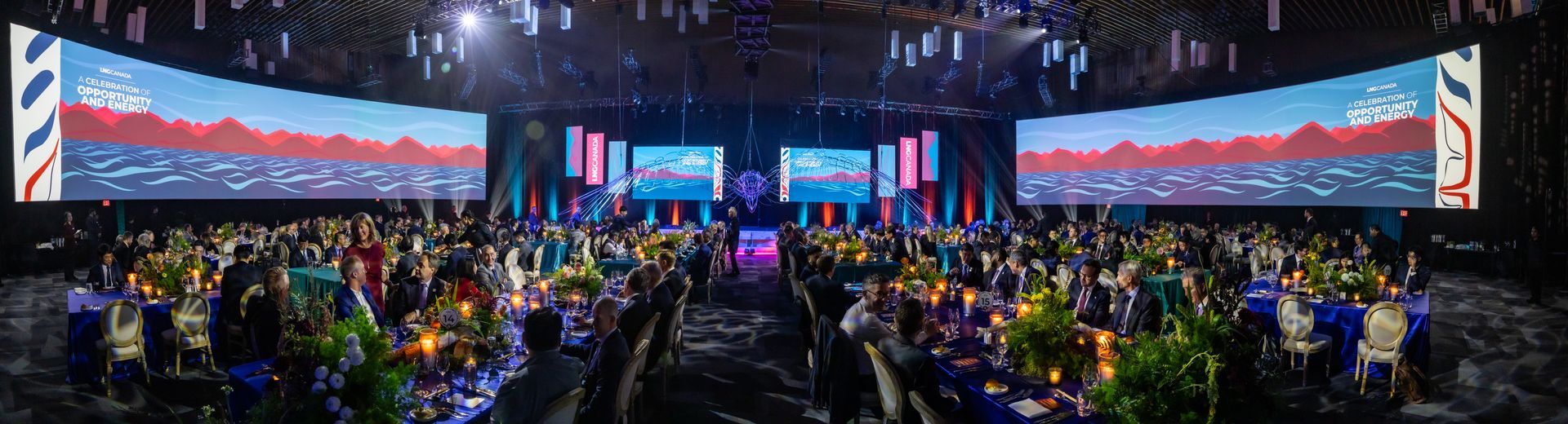 A large event hall with people seated at tables, large screens, and stage lighting.
