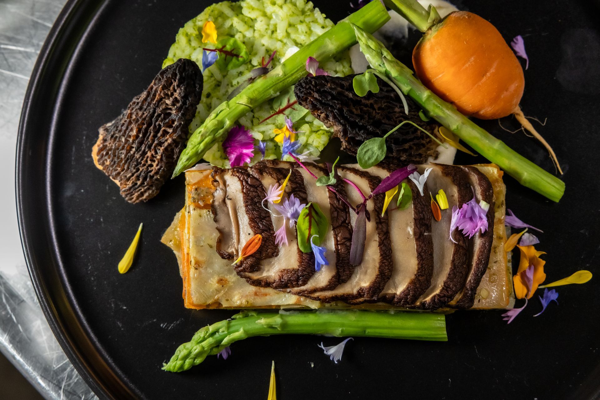 Close-up of a gourmet dish on a black plate: sliced mushrooms, asparagus, carrot, and green puree, garnished with flowers.