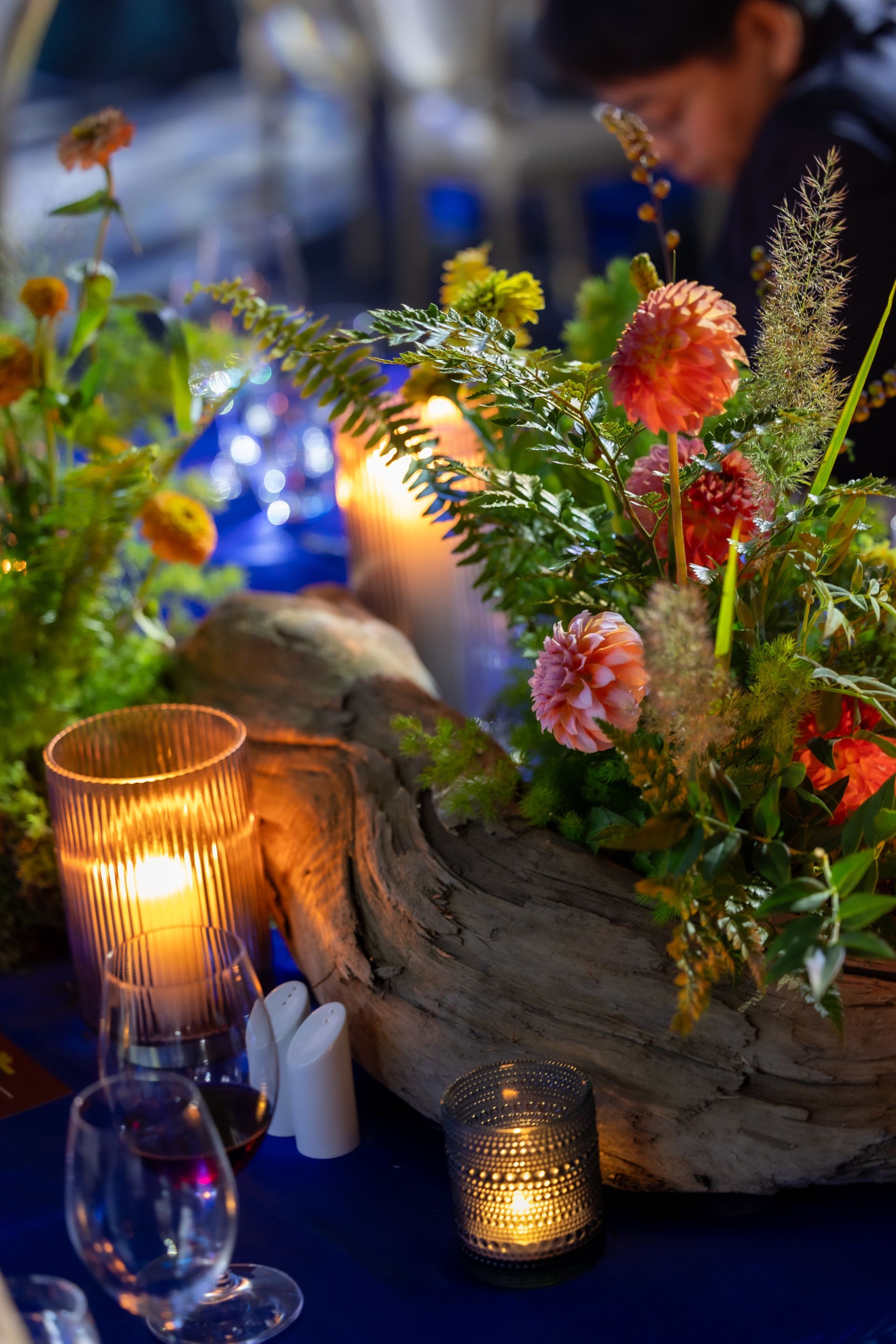 Table centerpiece with flowers, candles, and a driftwood base, on a blue tablecloth.