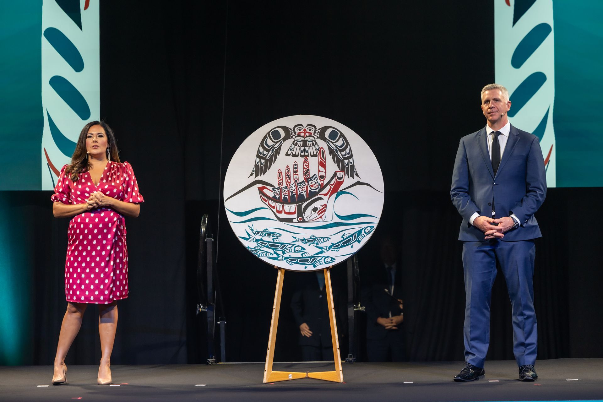Two people flanking a round artwork on an easel; the art features a boat with hands and a wave design.