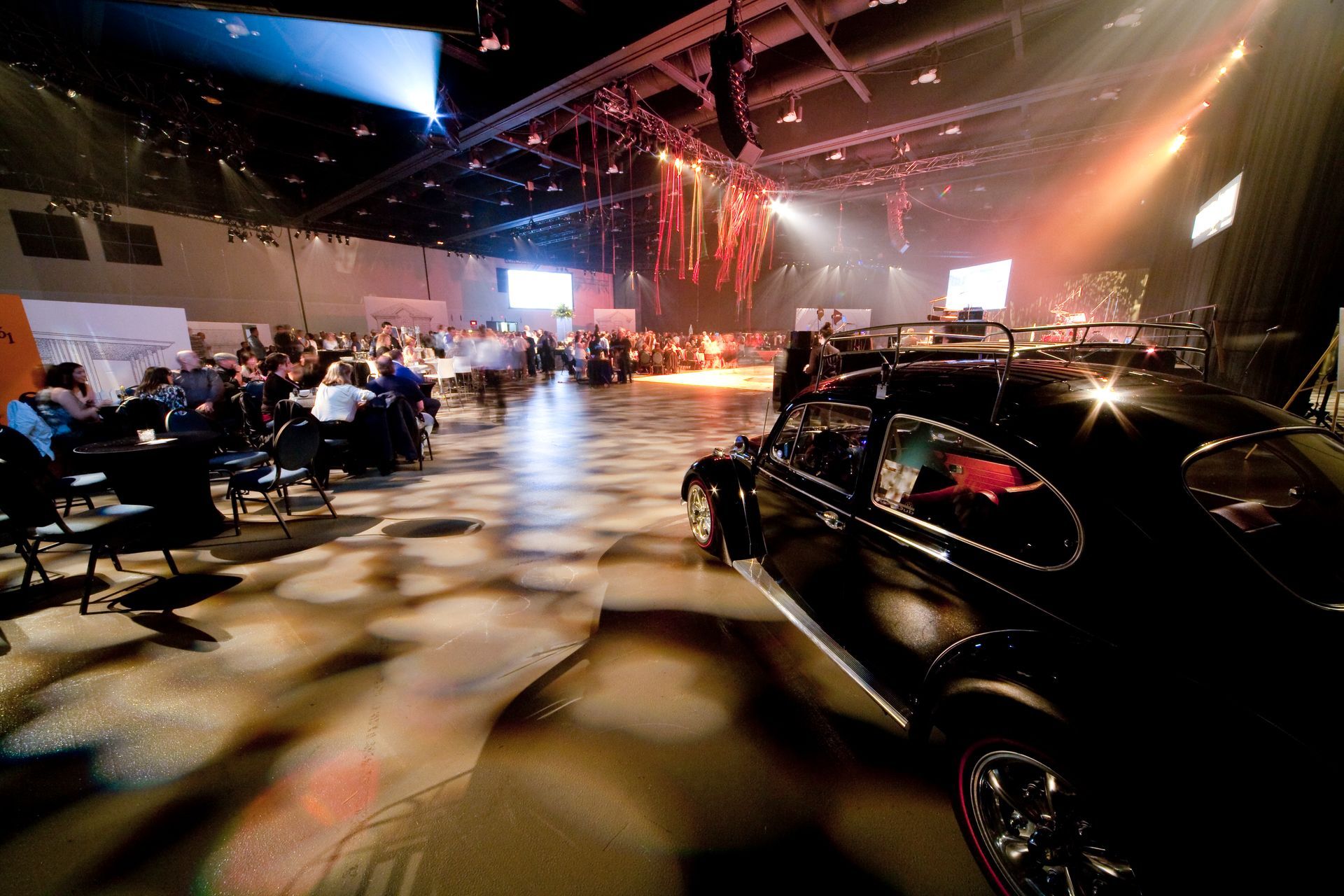 A black car is parked in a large room with people sitting at tables.
