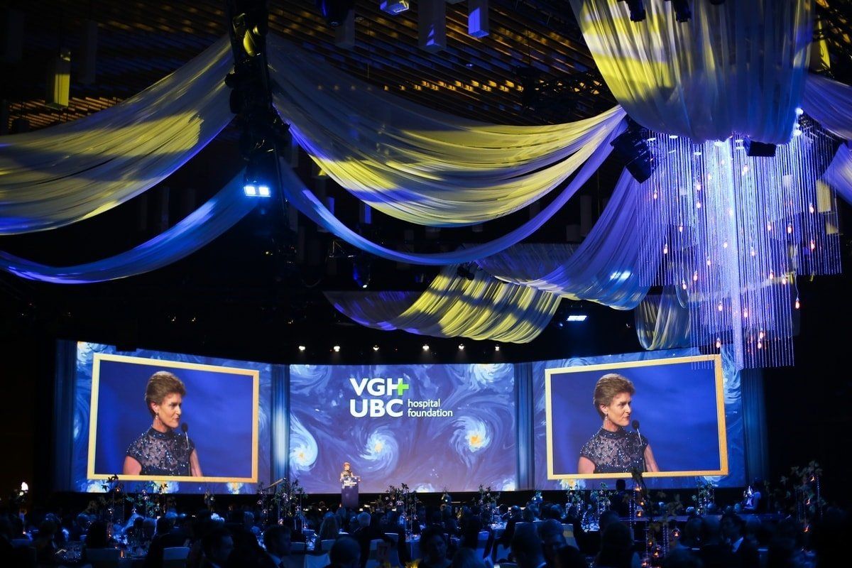 A woman is giving a speech in front of a large screen that says vgh ubc