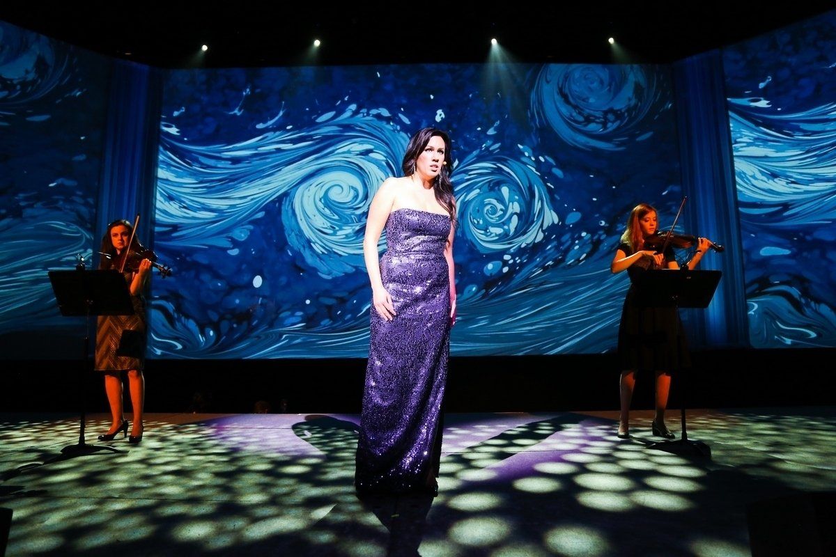A woman in a purple dress is singing on a stage with two women playing violins.