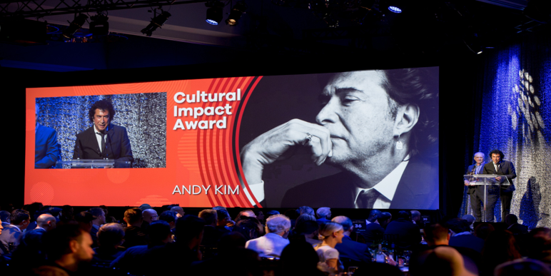 A man is giving a speech in front of a large screen that says cultural impact award.