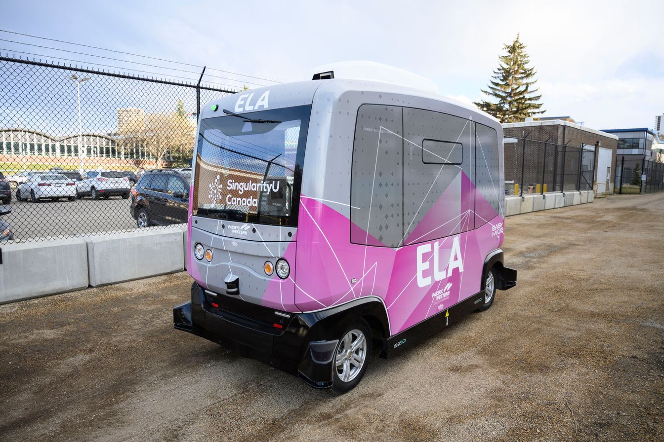 A pink and white bus is parked in a parking lot next to a chain link fence.