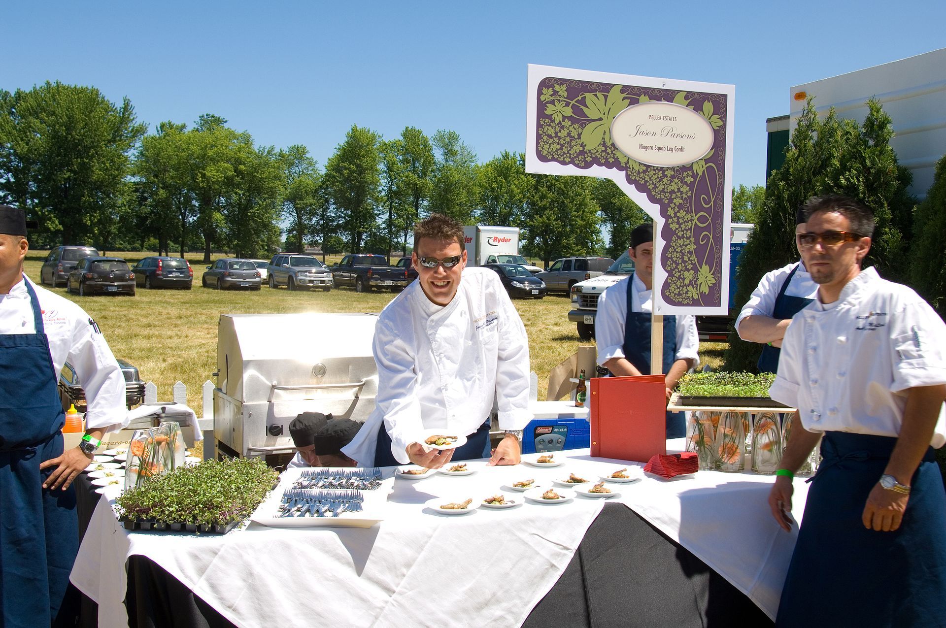 Chefs at an outdoor food stand, serving small plates under a sign. Bright sunny day with parked cars in the background.