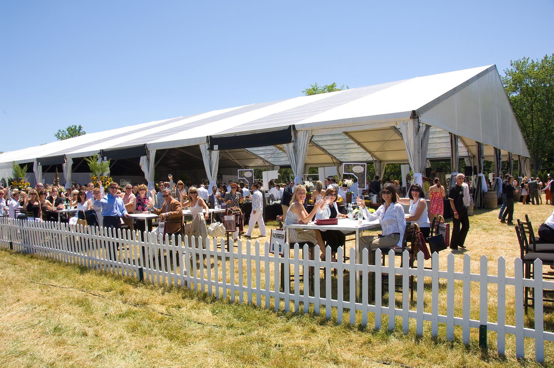 Large event tent with a crowd gathered behind a white picket fence on a sunny day.