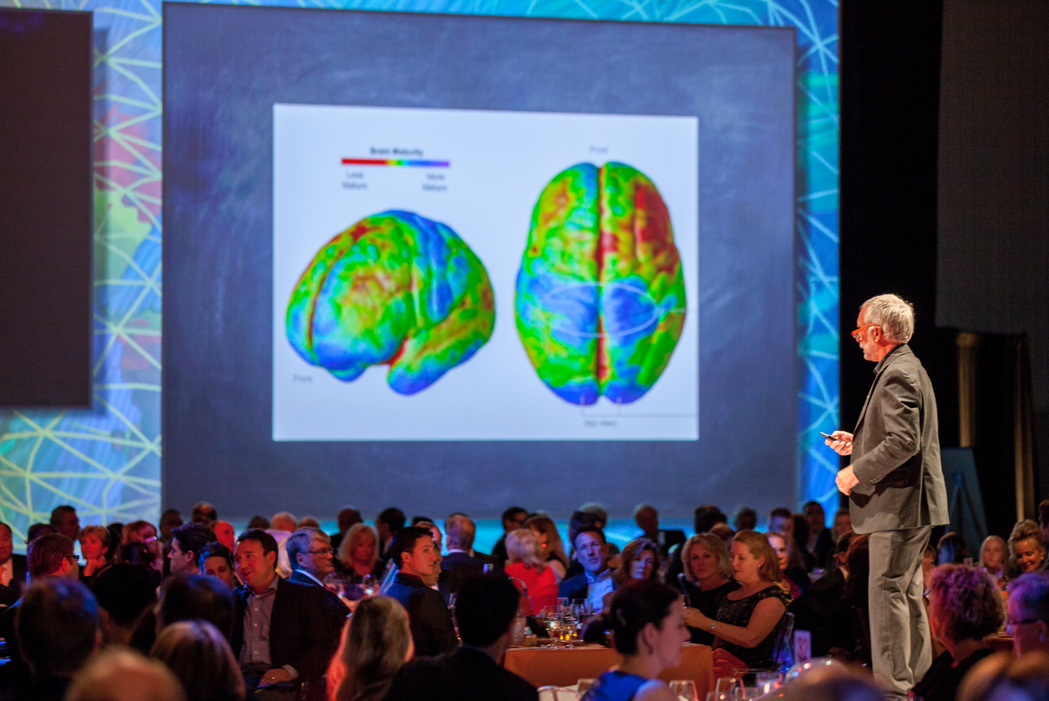 A man is giving a presentation in front of a large screen with a picture of a brain on it.
