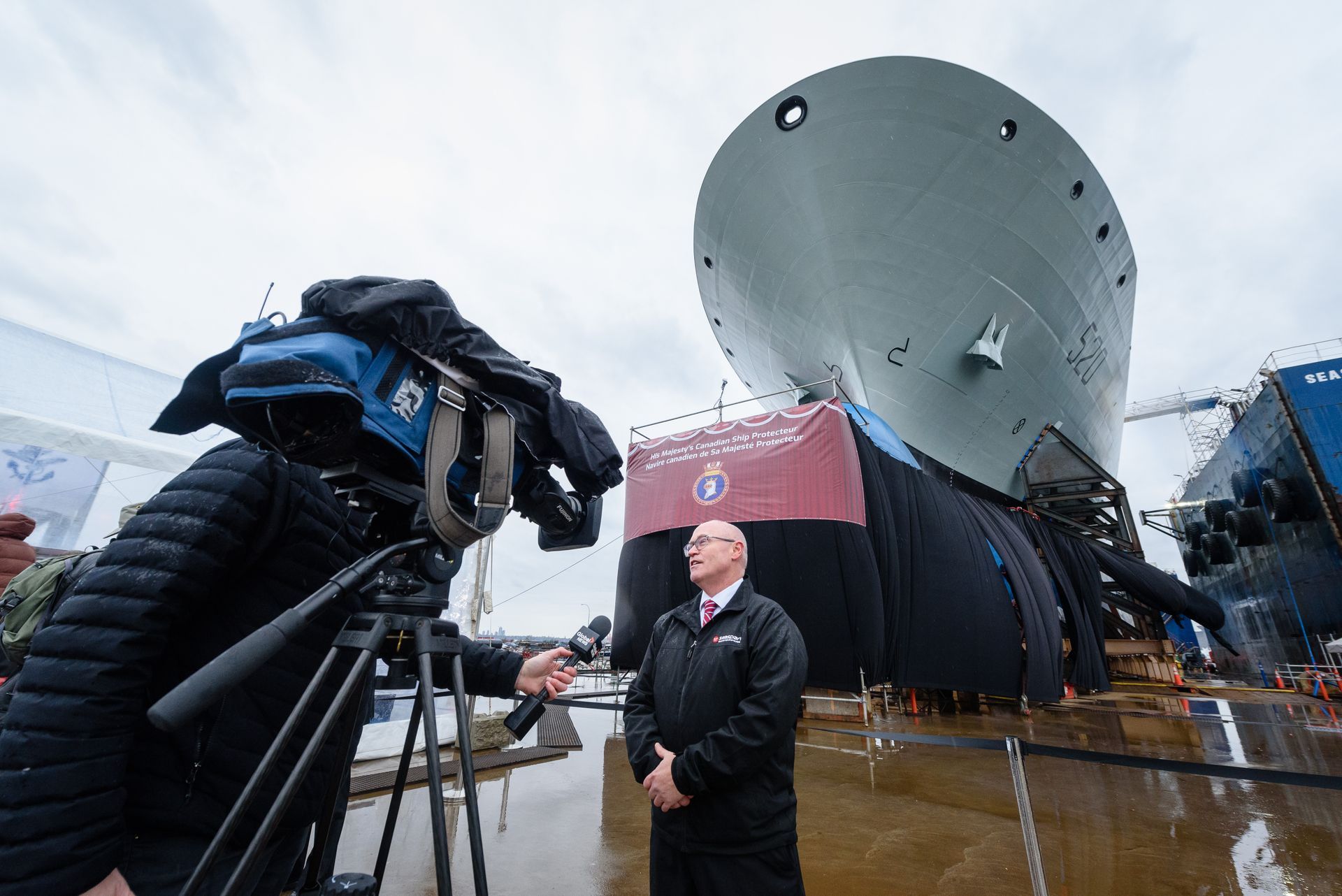 A man is standing in front of a camera in front of a large ship.