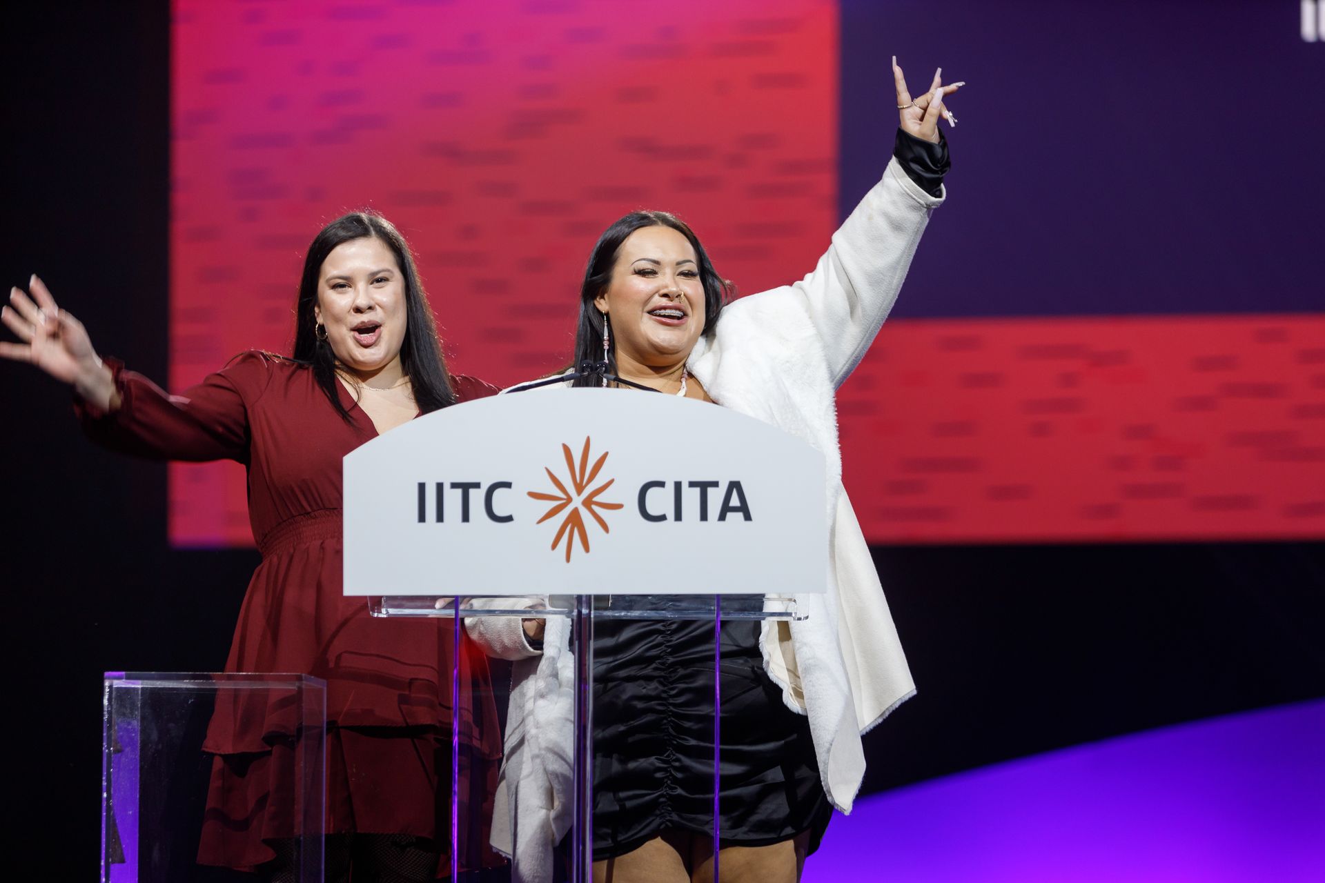 Two women on stage, smiling with arms raised, at IITC CITA event.