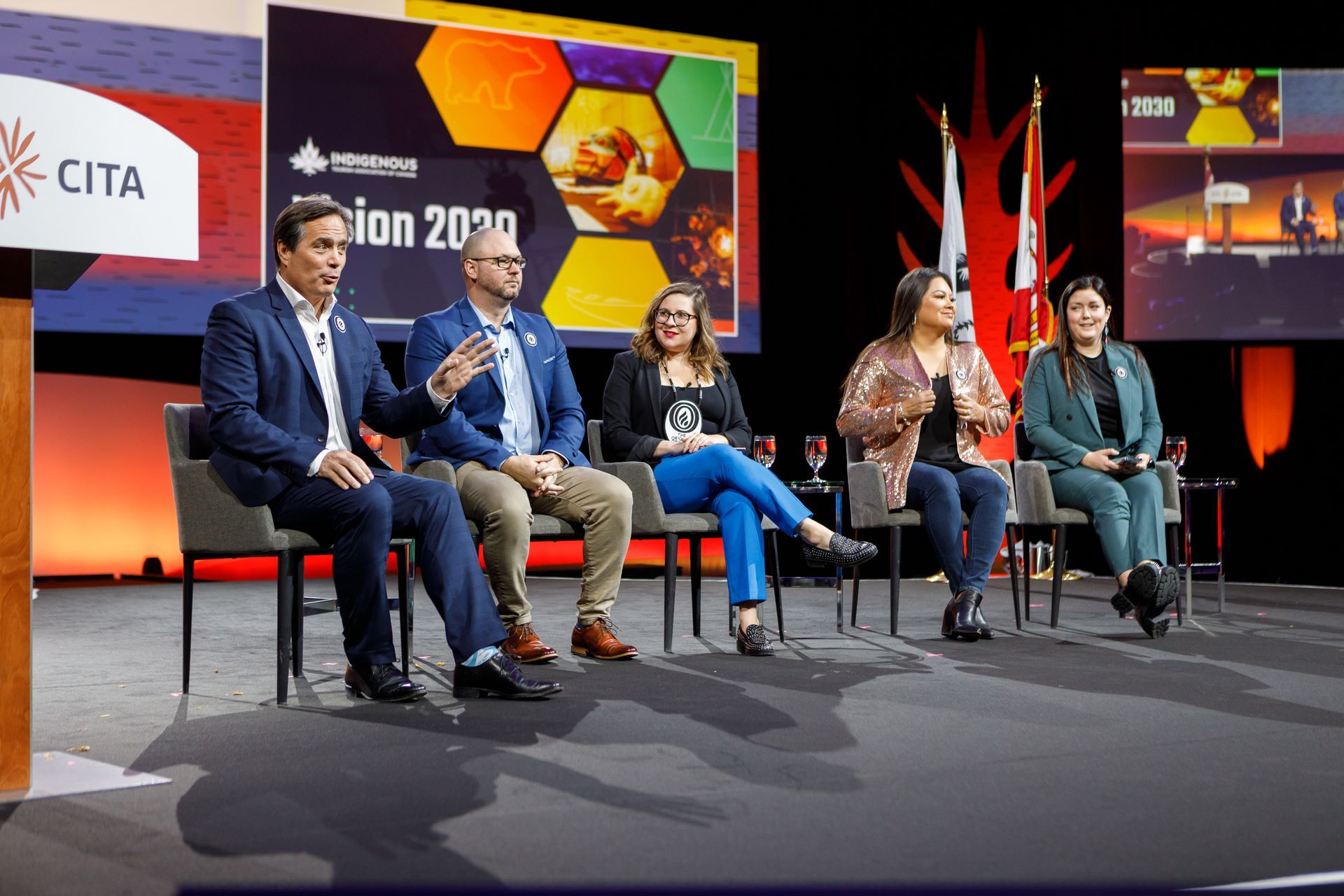 Panel of five people on stage at a conference, discussing; backdrop with logo, stage lights.