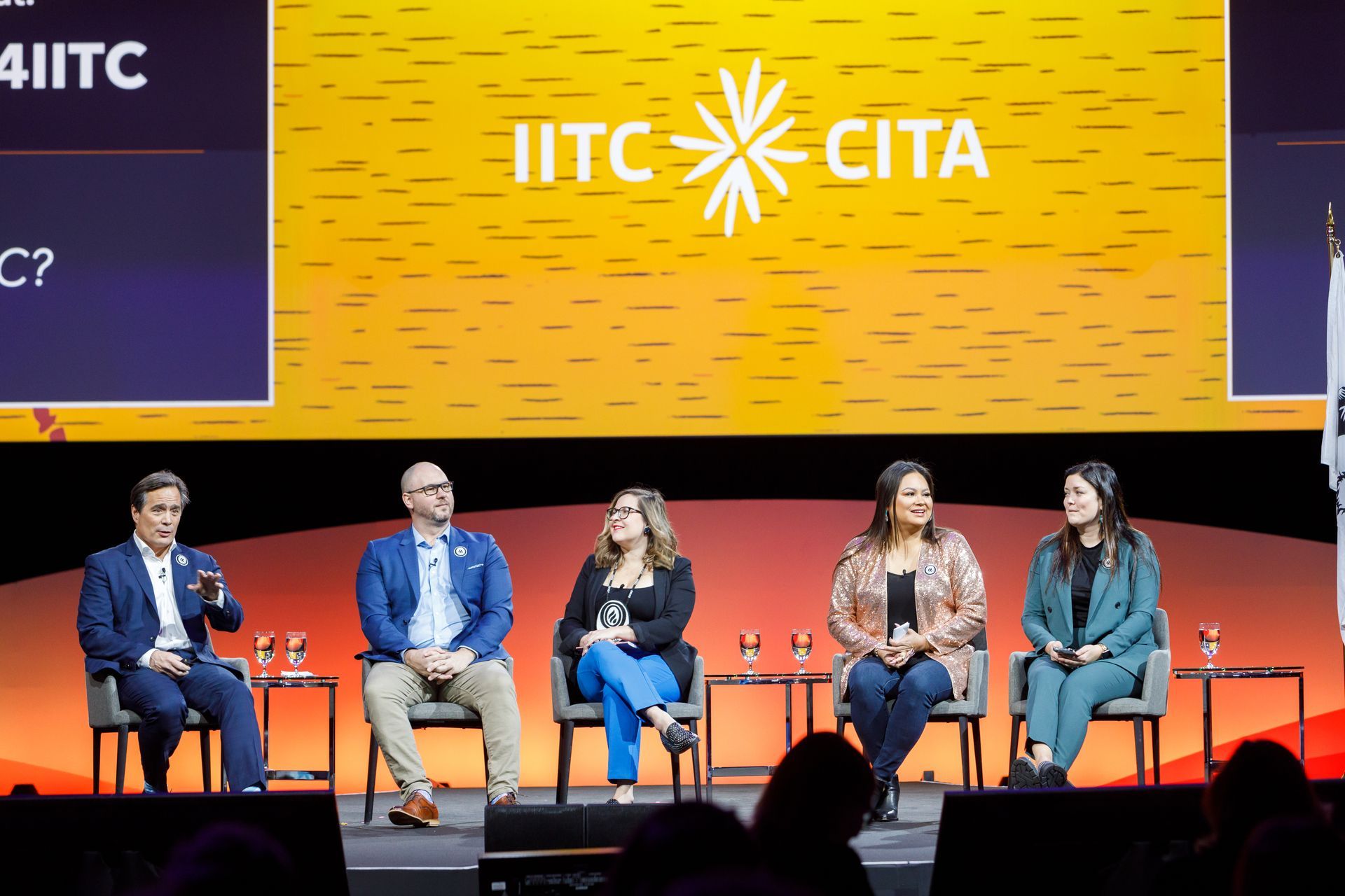 Panel of five people on stage, discussing at IITC conference. Bright stage backdrop.