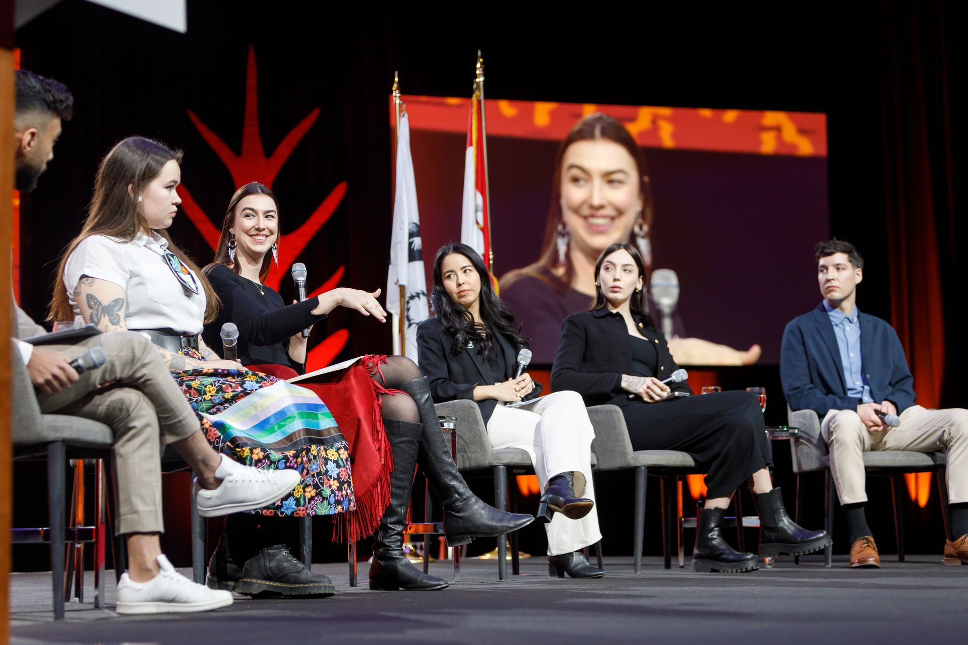 Panel of people seated on stage, flags behind them, woman's face on screen.