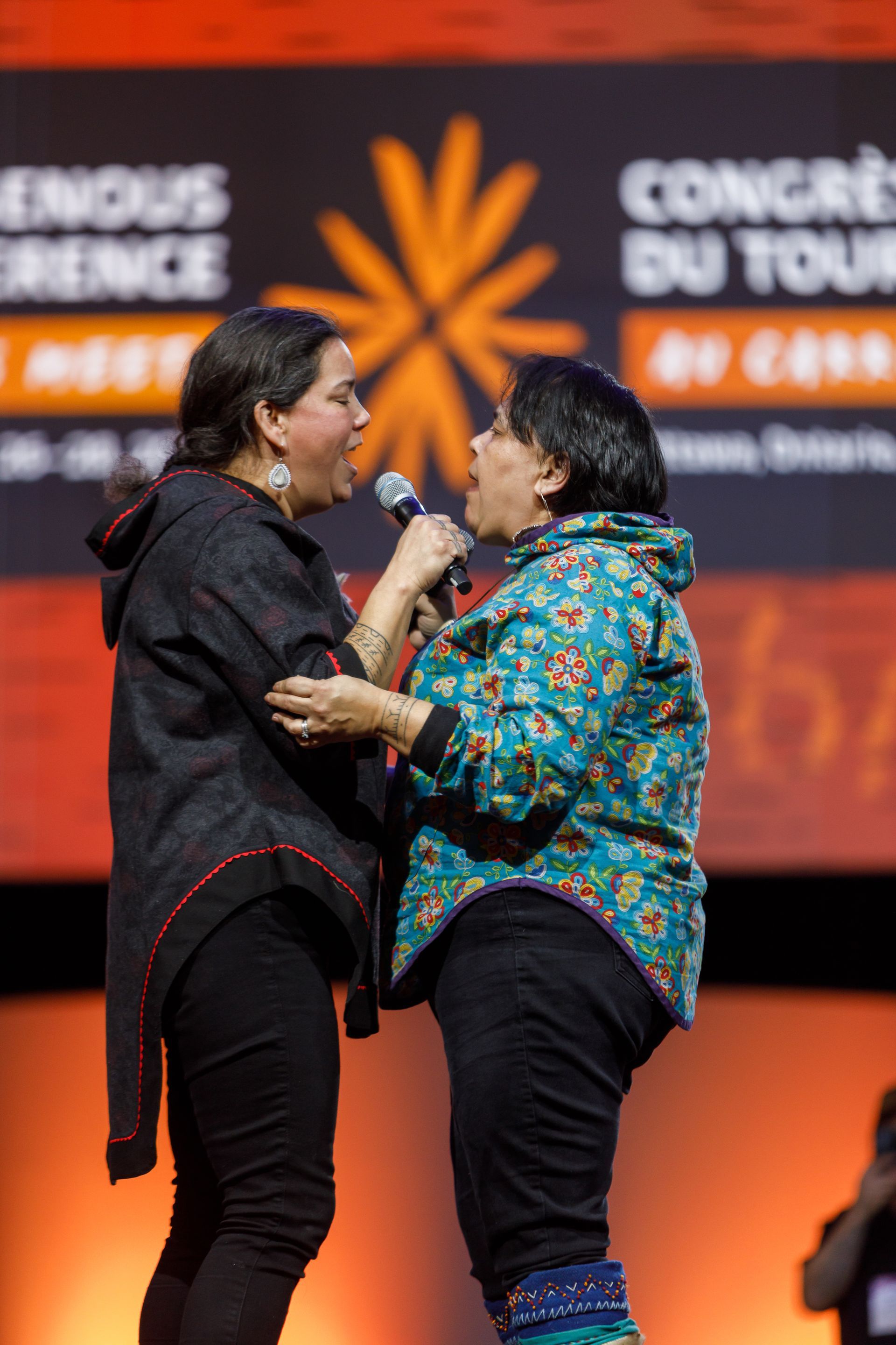 Two women on stage, one holding a microphone, both singing. Conference backdrop, orange and white.