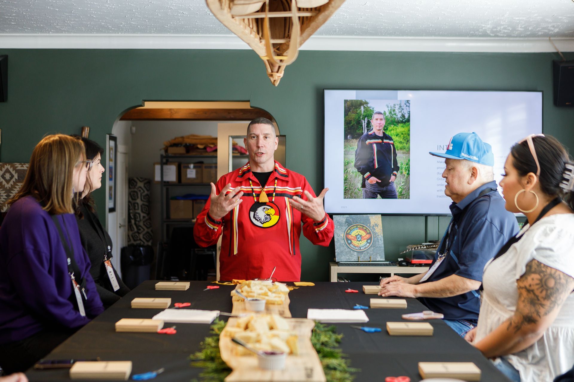 A man in a red shirt is giving a presentation to a group of people sitting around a table.