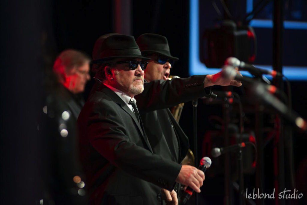 Two men in suits and hats are singing into microphones on a stage.