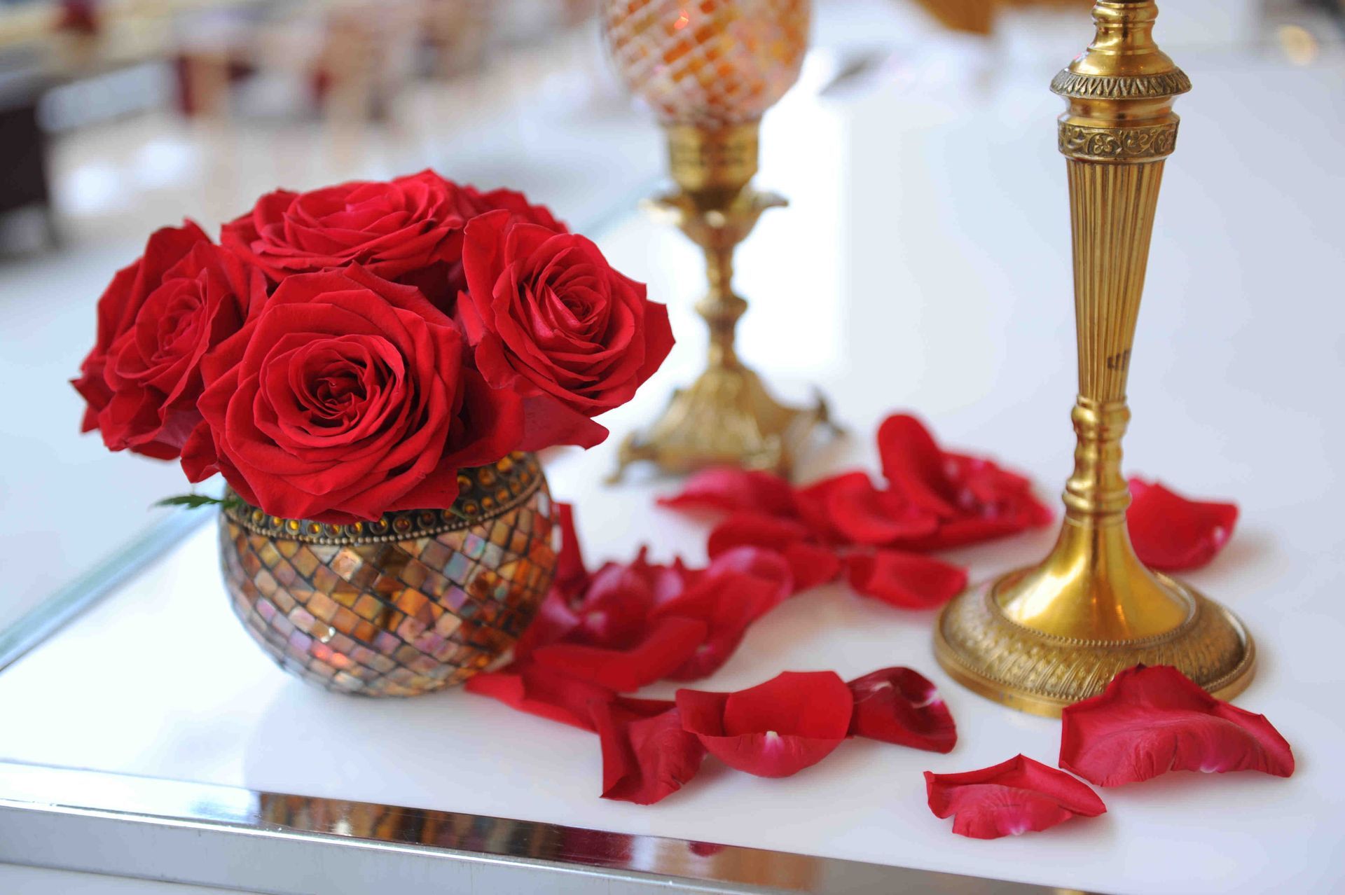 A vase of red roses sits on a table next to candles and rose petals