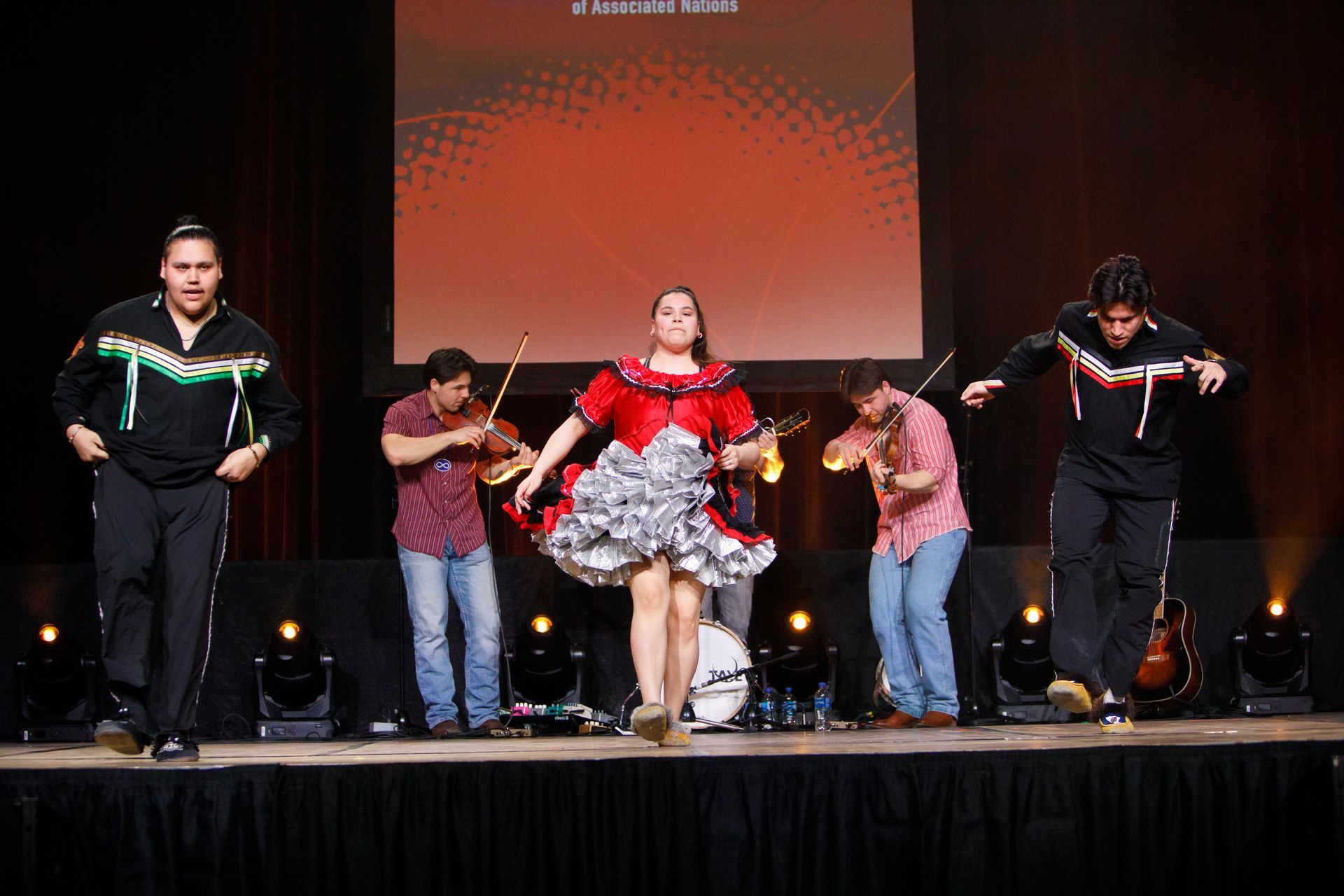 A group of people are dancing on a stage in front of a large screen.