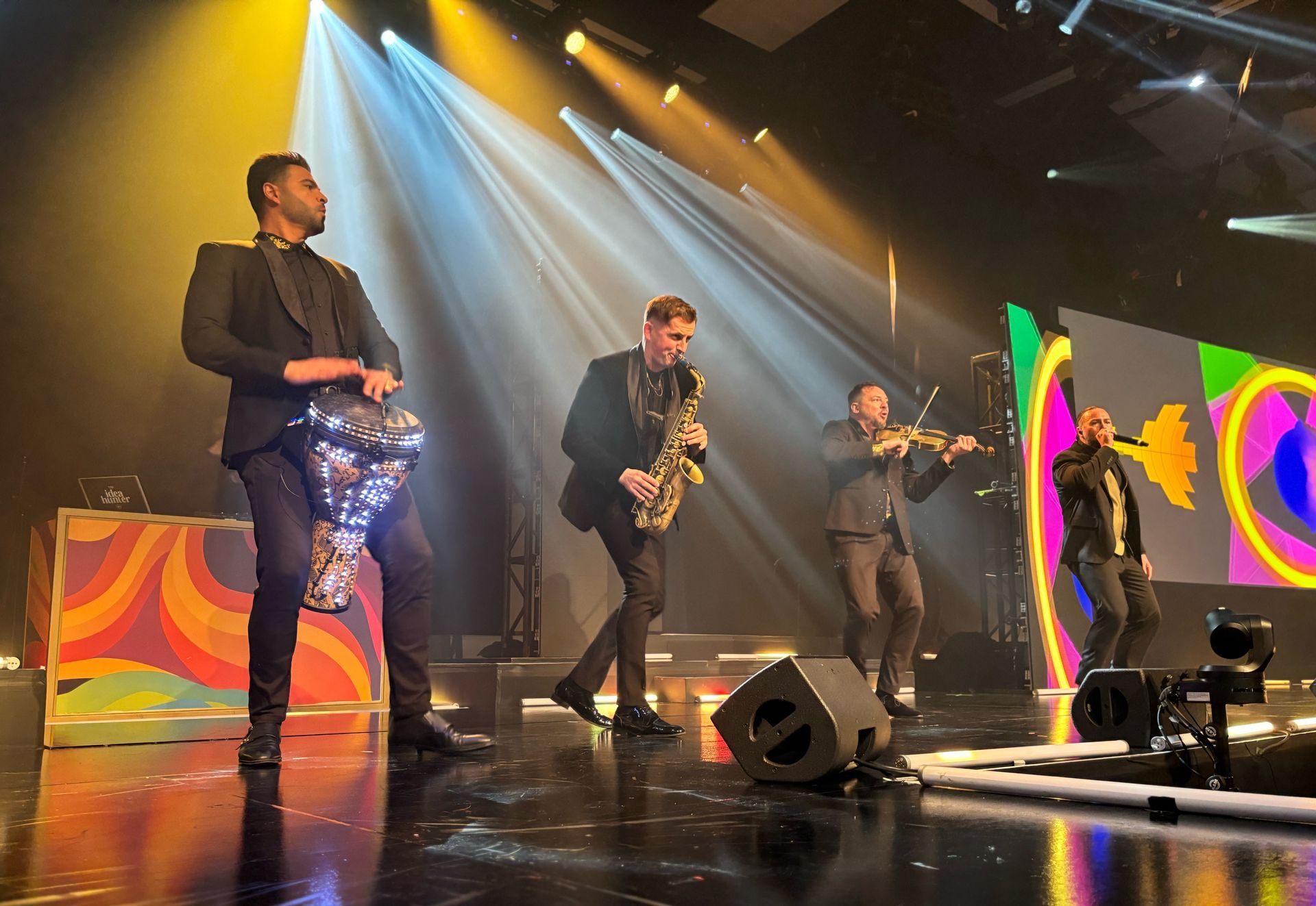 A band performs on stage. Musicians play saxophone, violin, and drums, under stage lights with a colorful backdrop.