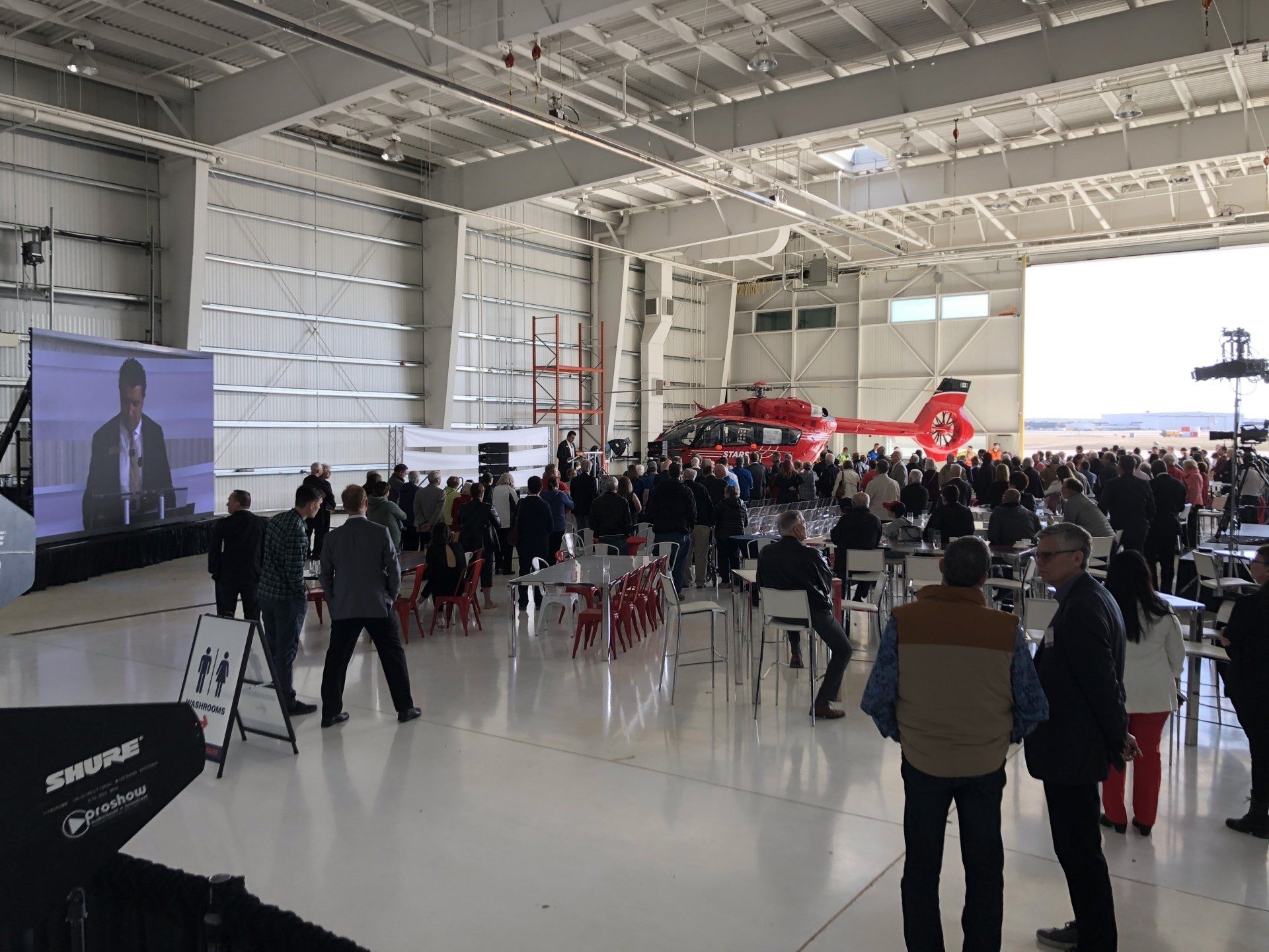 A large group of people are gathered in a hangar with a helicopter in the background.