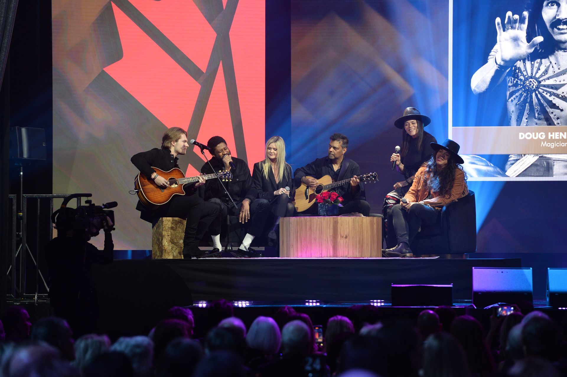 A group of people are sitting on a stage playing guitars and singing into microphones.