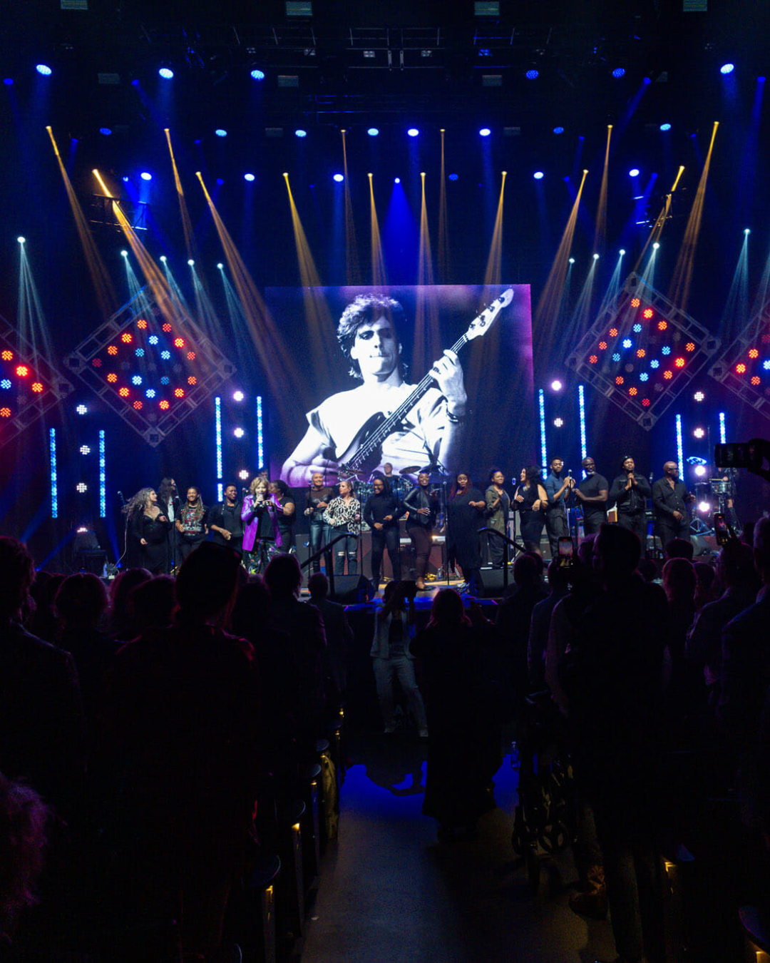 A group of people are standing in front of a large screen with a picture of a man playing a guitar on it