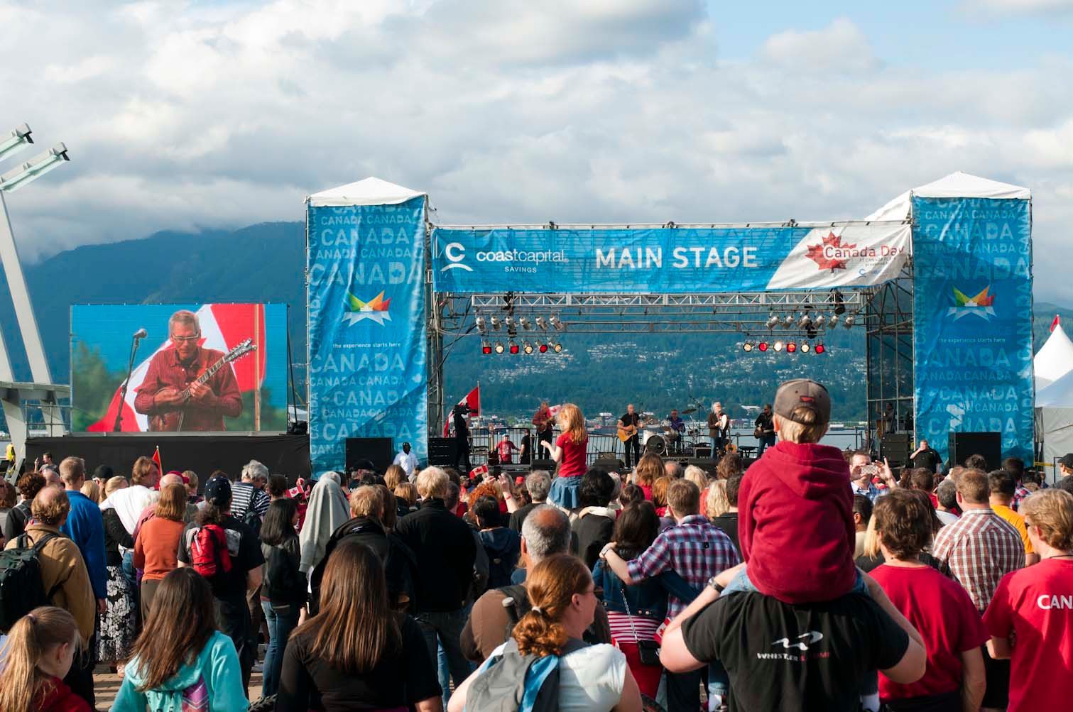 Large crowd at outdoor concert; stage with musicians, giant screen shows performer, blue sky.