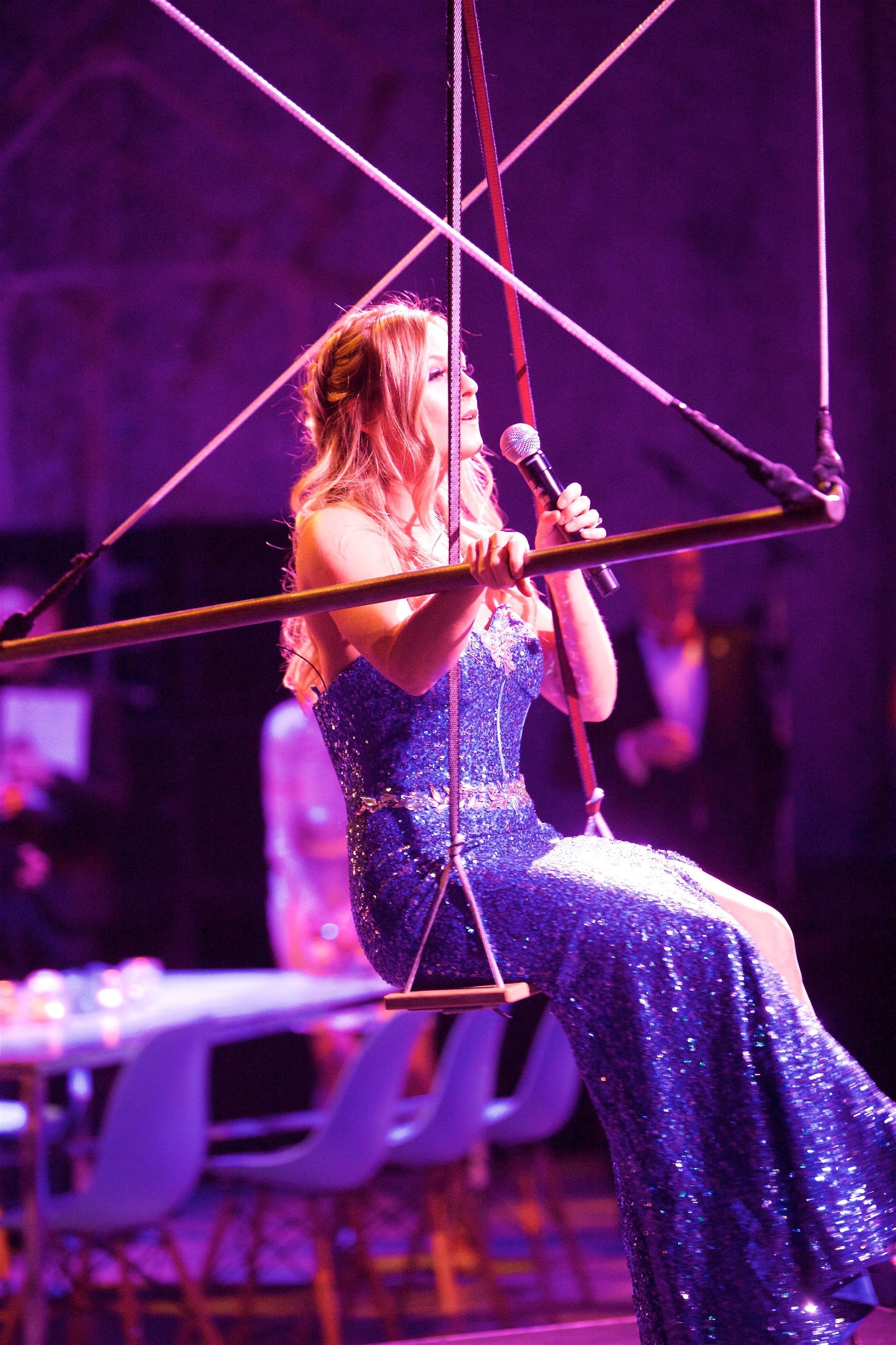 A woman in a blue dress is singing into a microphone while sitting on a swing