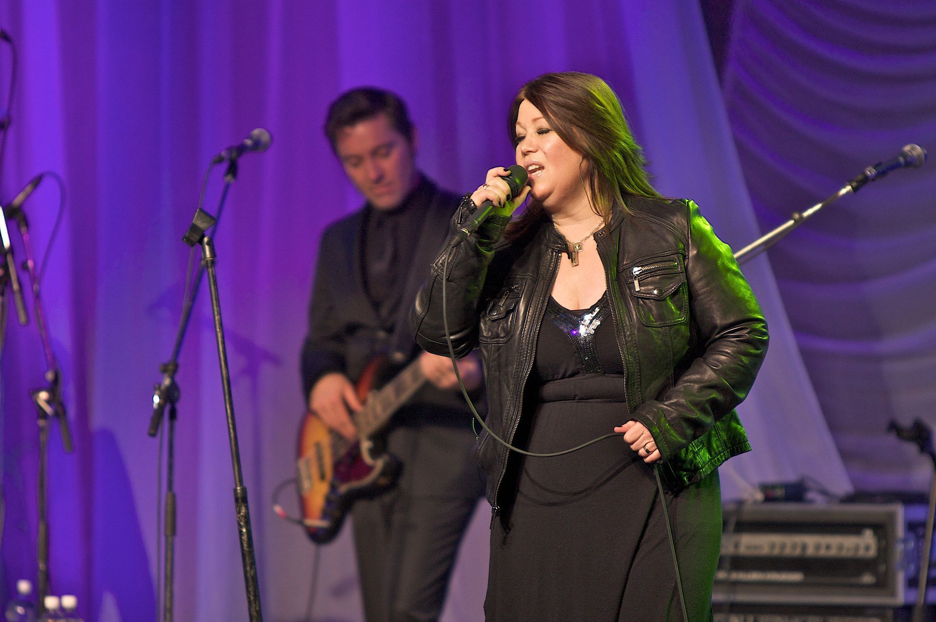 A woman singing into a microphone with a man playing a guitar in the background.