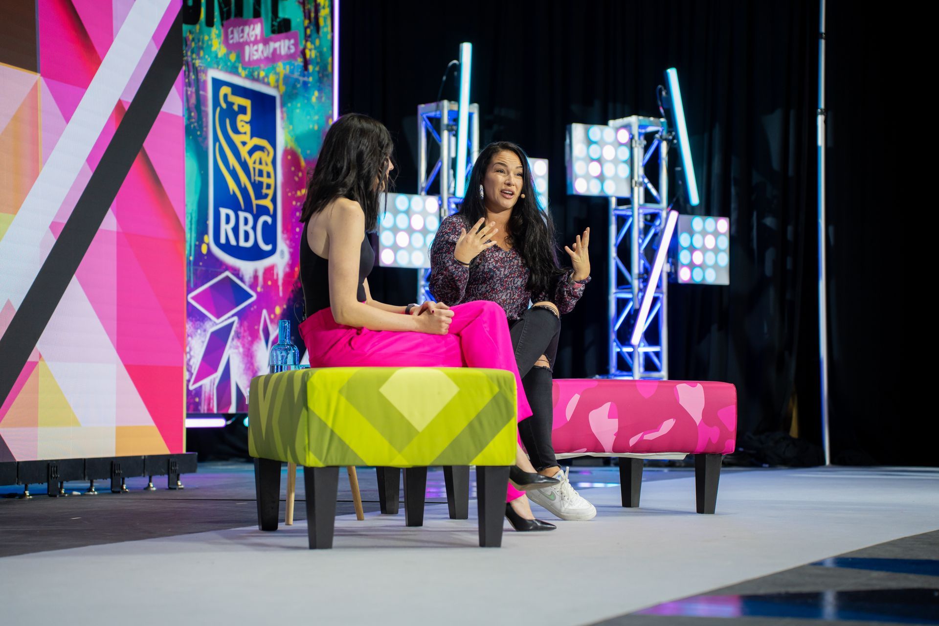 Two women are sitting on ottomans on a stage talking to each other.