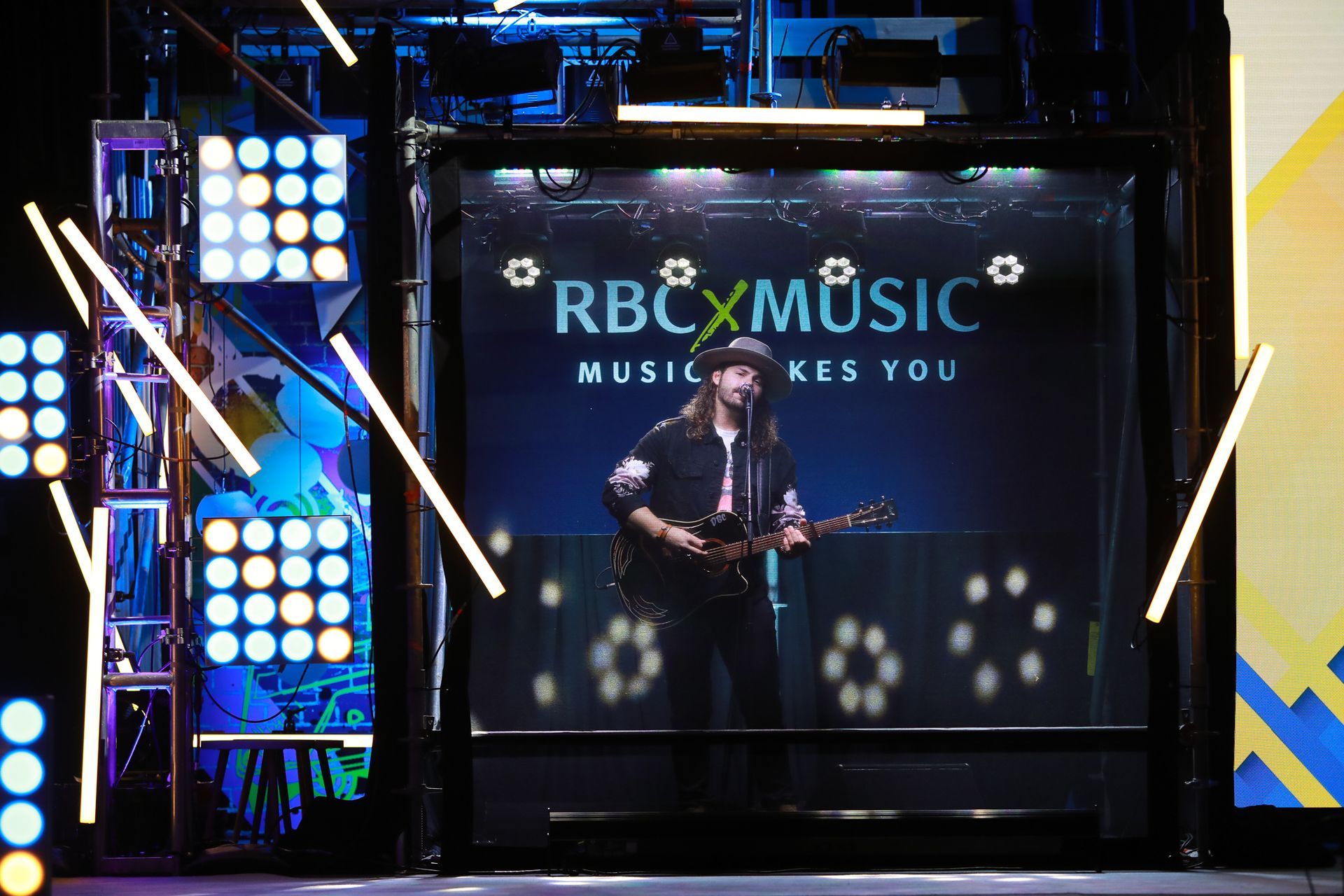 A man is playing a guitar in front of a screen that says rbcxmusic