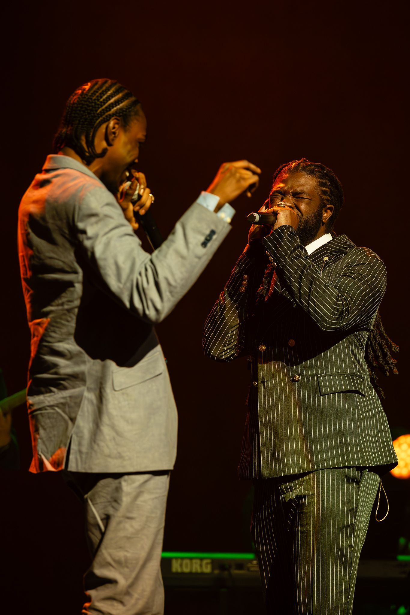 Two men on stage, singing into microphones. One in a gray suit, the other in a black patterned suit.