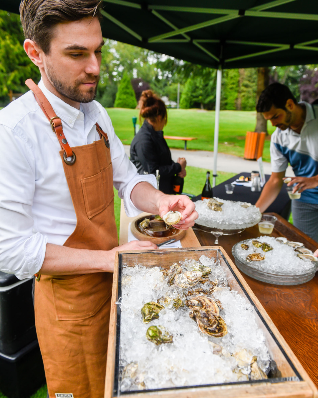 A man in an apron is holding a tray of oysters on ice.
