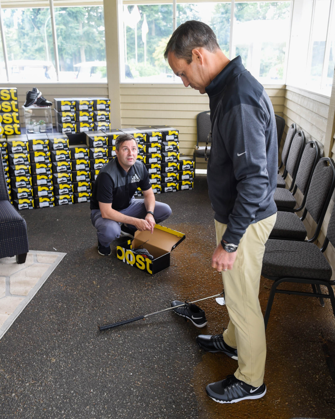 Two men are looking at a golf club in front of boxes that say cost