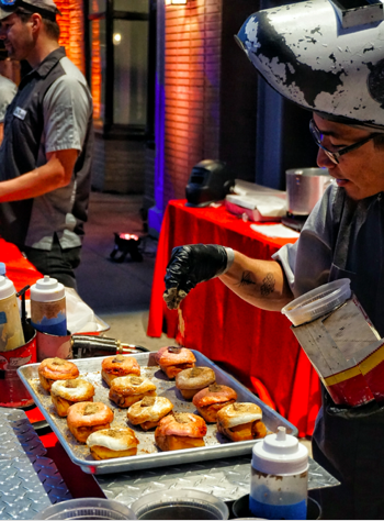 A man wearing a welding helmet is pouring sauce on a tray of donuts