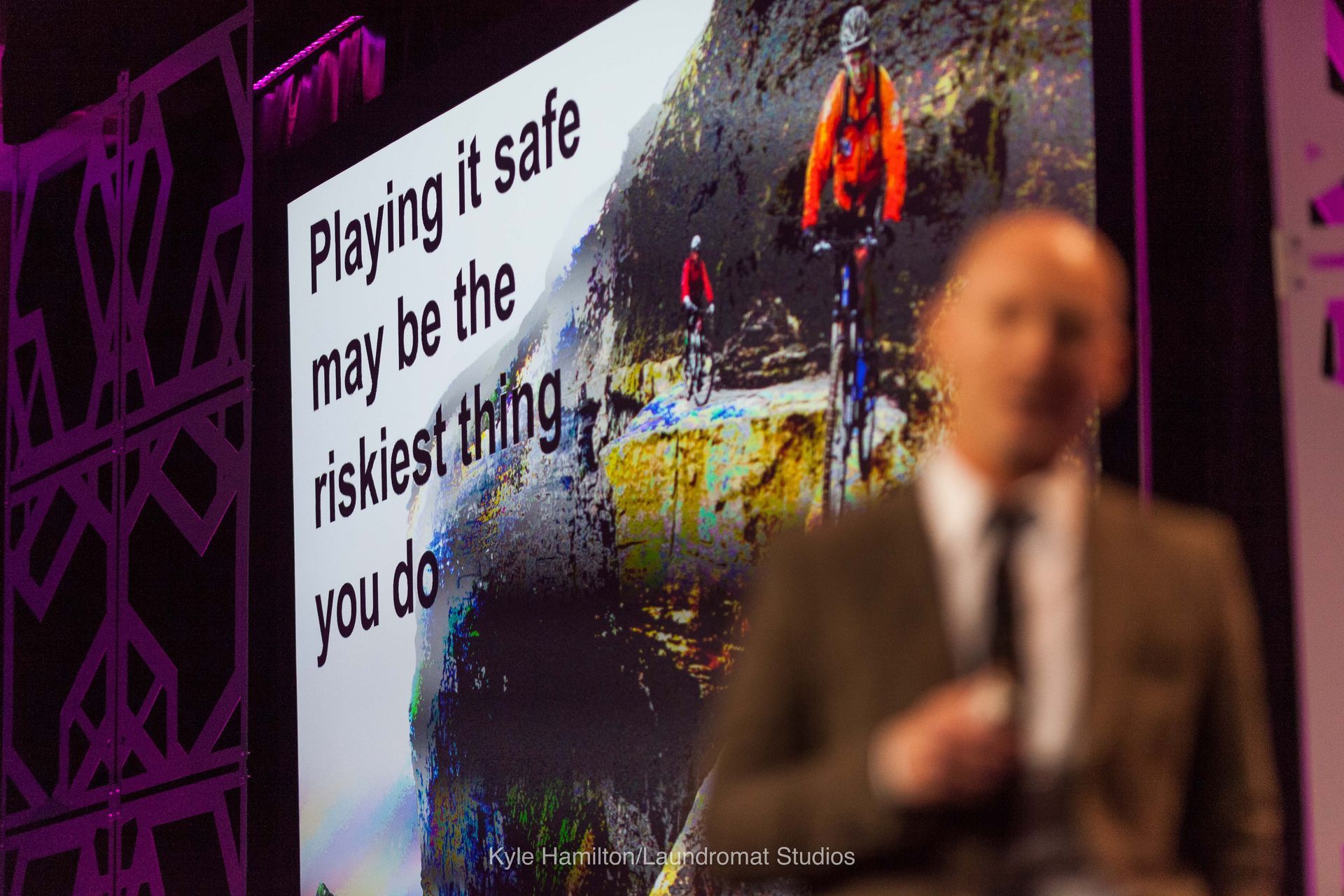 A man is standing in front of a screen that says playing it safe may be the riskiest thing you do