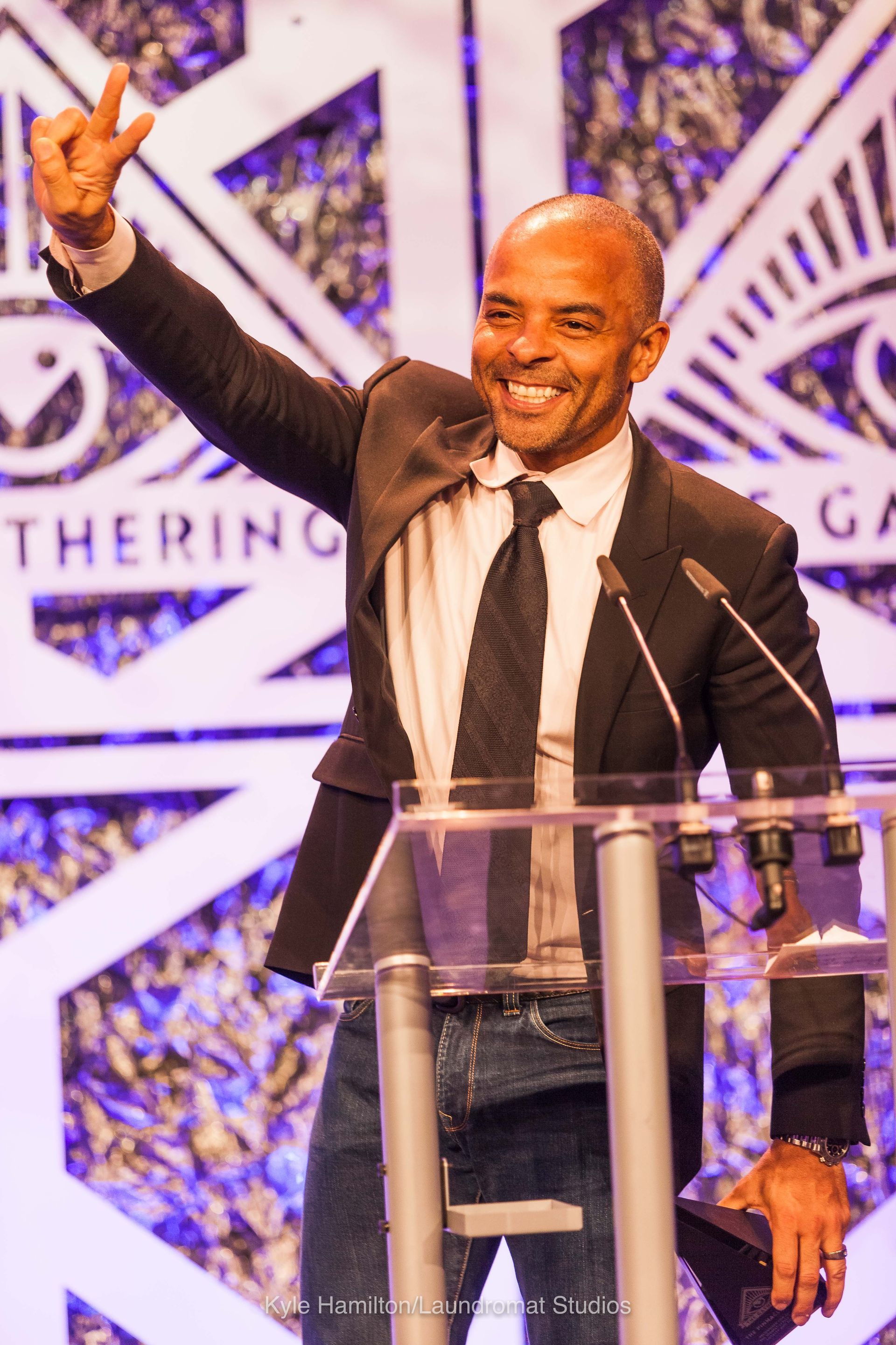 A man in a suit and tie stands at a podium giving a peace sign