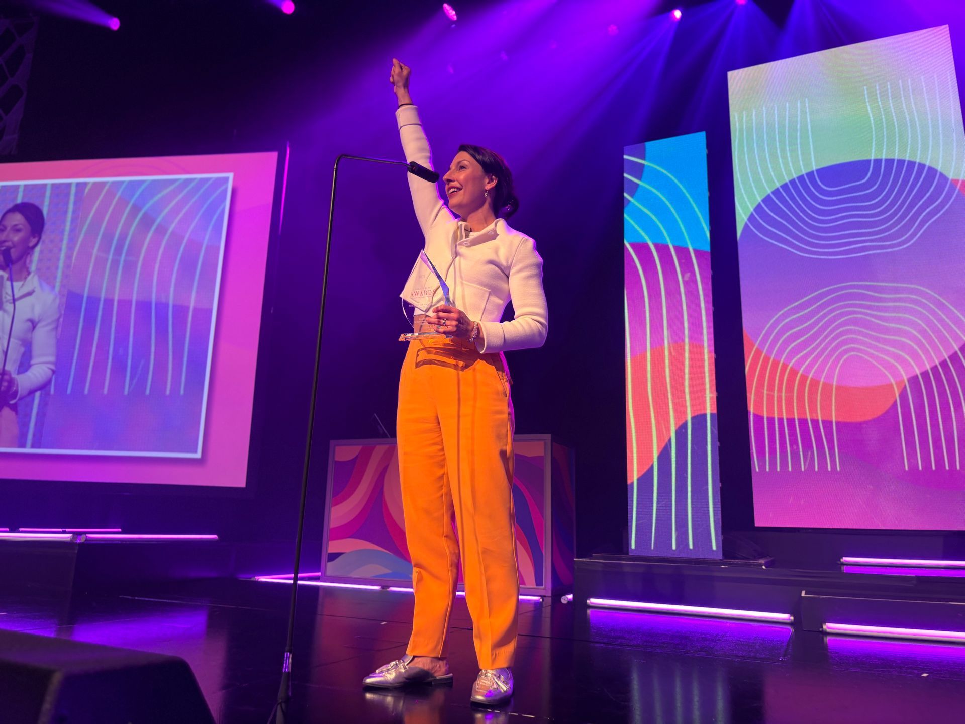 Woman on stage, arm raised, wearing orange pants and white jacket, performing with colorful backdrop.