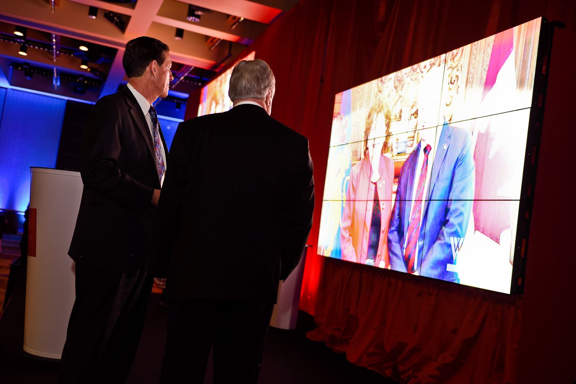 Two men are looking at a large screen with a picture of a man and woman on it.